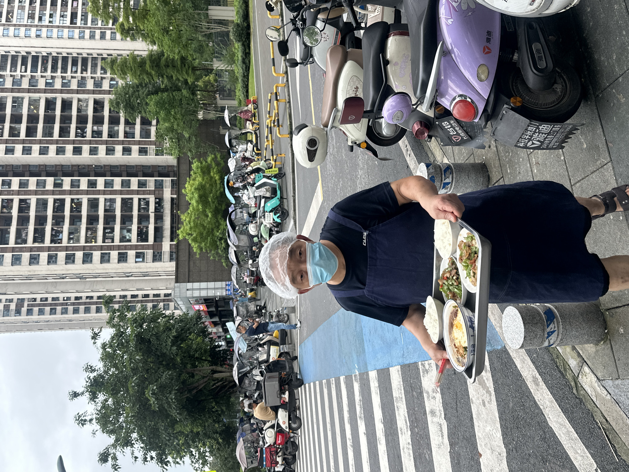 In Chongqing High-tech Zone, China, a person wearing a blue apron and a hairnet is holding a tray of food. The tray contains several dishes, including rice, a stir-fry with vegetables and meat, and another dish with a fried egg over rice. The person is standing on a sidewalk next to a street lined with parked electric scooters. The scooters are of various colors, including white, purple, and green, and many are equipped with storage boxes.
In the background, tall, modern buildings are visible, suggesting an urban environment. Trees are also present, adding a touch of greenery. The lighting indicates it is daytime, and the sky appears overcast, suggesting cloudy weather. The presence of many scooters suggests this may be an area where delivery services are prevalent. The activity appears to be related to food delivery or service, with the individual carrying a prepared meal. Some scooters have license plates with Chinese text, and one scooter has visible branding for "Yadea" (雅迪), a well-known electric vehicle manufacturer in China. Other visible text includes information about a "Technology College" and a "24-hour free door-to-door service" with a phone number.