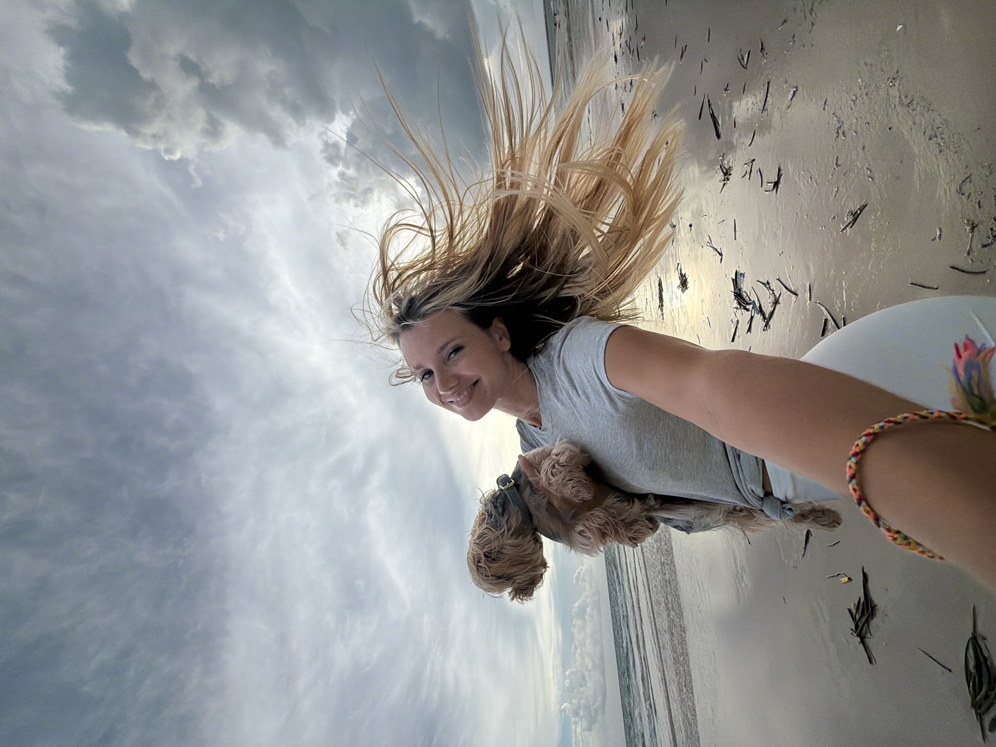 A selfie-style photograph captures a young blonde woman holding a small, light-brown dog at a beach. The woman is wearing a grey t-shirt and white pants, and a colorful braided bracelet is visible on her left wrist. Her blonde hair is blowing dramatically in the wind, and she is smiling at the camera. The dog, held in her left arm, has its head turned away from the camera. The background shows a beach with wet sand, scattered debris, and the ocean meeting the shore under a cloudy sky. The location is identified as Sakhu, Thailand, though this specific detail is not visually evident in the image itself.