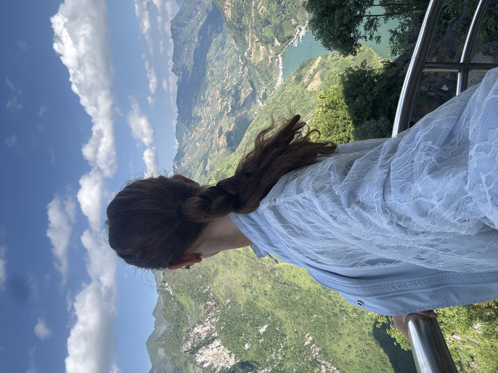 A woman with brown hair braided back is seen from behind, looking out at a mountainous landscape. She wears a light blue, lacy jacket and holds onto a metal railing. The background features a valley with green mountains and a body of water. The sky above is blue with white clouds. The location is identified as Đồng Văn Commune, Vietnam, based on the landscape features.