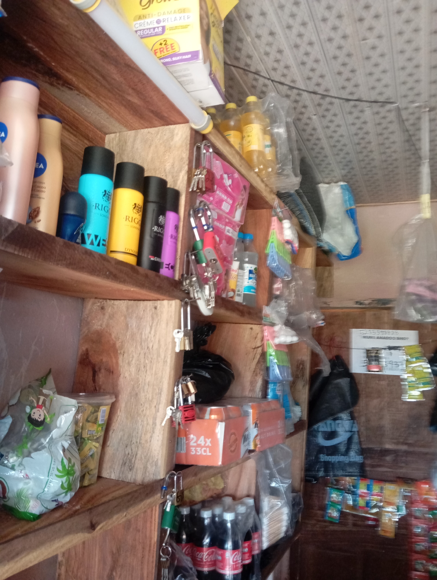 The image depicts the interior of a small shop, likely a convenience store, located in Jalingo, Nigeria. The shop is characterized by wooden shelving filled with various products. On the top shelf, there are bottles of hair relaxer, and several spray cans including RIGOROUS brand in blue, yellow, black, and purple. Below these, on the next shelf, are bottles of what appears to be beverages, including Coca-Cola. Also visible are bags of snacks, packaged goods, and an assortment of keys and padlocks hanging from hooks. The lighting appears to be artificial, suggesting an indoor setting. The overall impression is of a modest retail space stocked with everyday consumer goods.