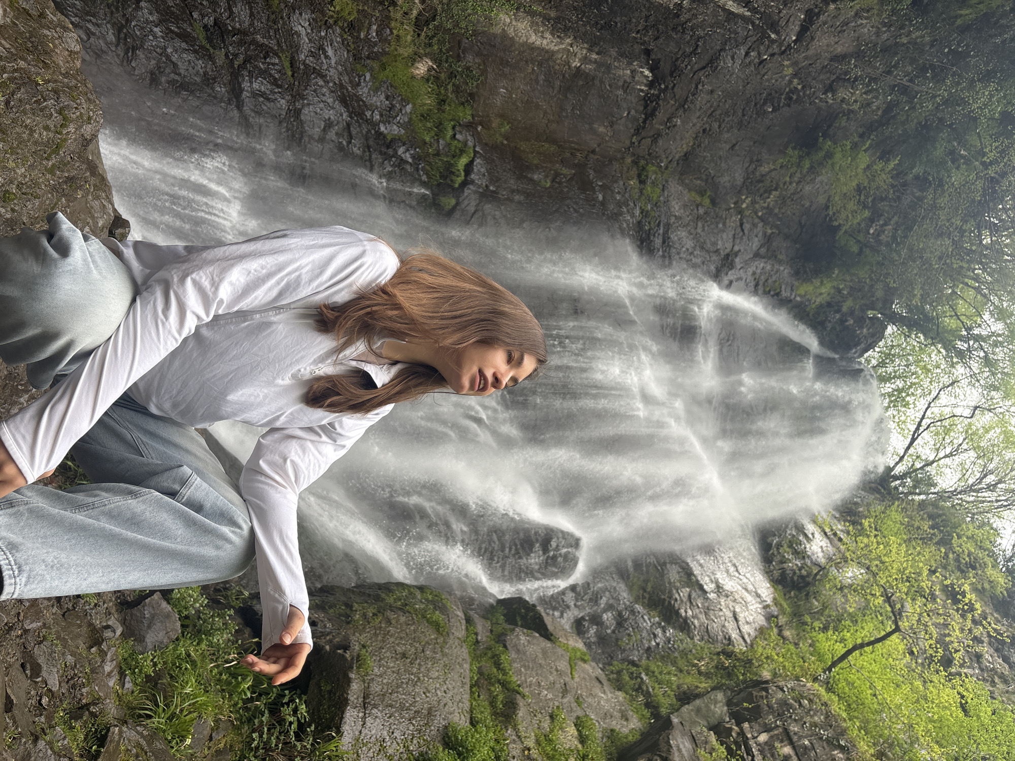 A woman is photographed in front of the Makhuntseti Waterfall, located in Kveda Makhuntseti, Georgia. The woman has brown hair and is wearing a white long-sleeved shirt and light blue jeans. She is positioned on rocks, looking downward towards the waterfall. The waterfall itself is cascading down a rocky cliff face, with some greenery visible on the rocks surrounding the waterfall. The scene is set in a natural environment.