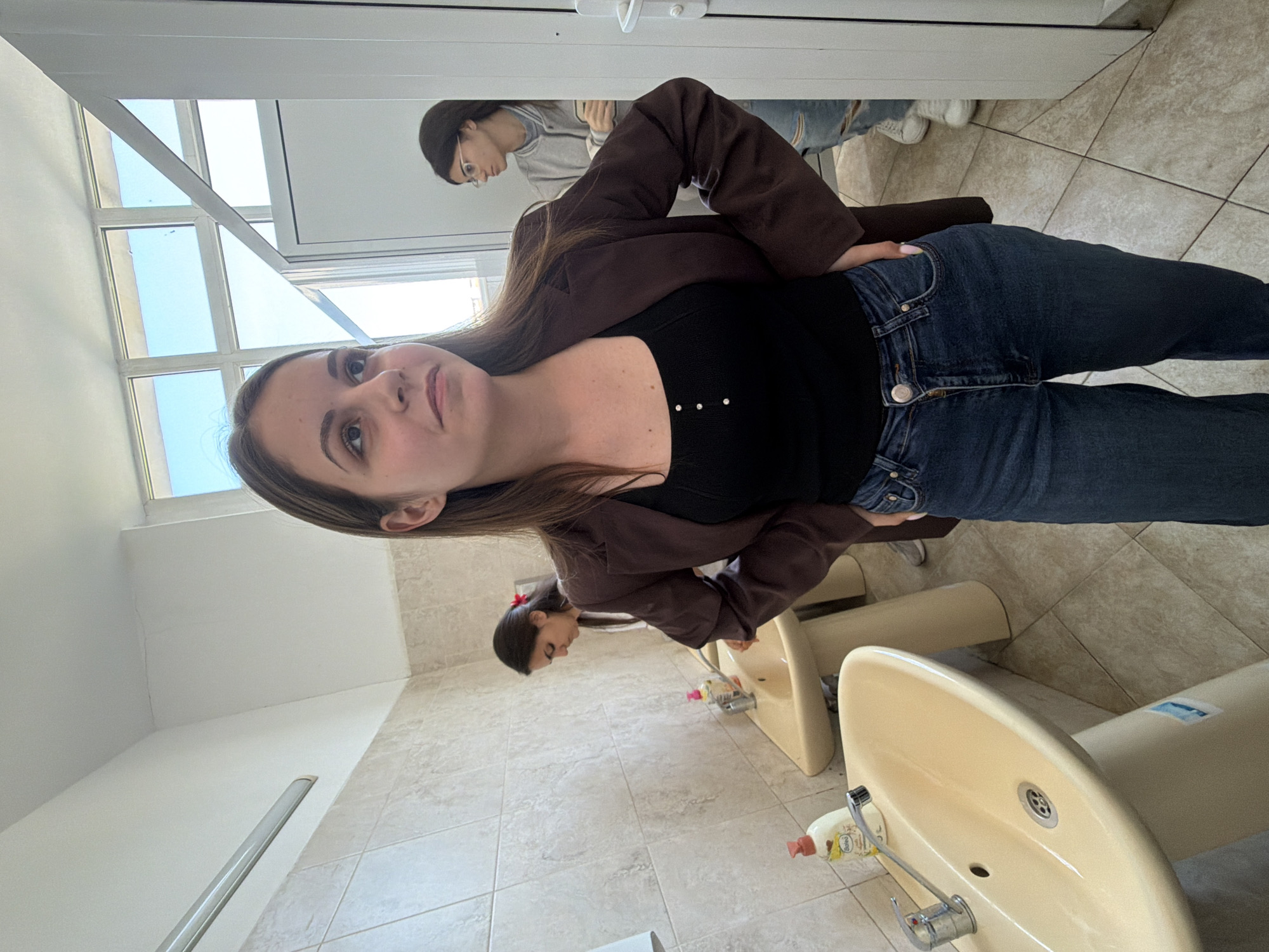 The image shows a brightly lit restroom with beige tiled walls and floors. Three young women are present. One woman, centered in the foreground, is wearing dark blue jeans, a black top, and a brown jacket. Her hand rests on her hip. Behind her to the right, another woman wearing glasses is standing with her arms crossed, wearing a grey sweatshirt and jeans. To the left and behind, a third woman with a red hair accessory is using one of the beige sinks. The room features several beige sinks and toilets. On one of the sinks there is a bottle of hand soap. Based on the architecture and fixtures, it is possible that the restroom is located in a public building or institution. No specific landmarks indicate the city of Burgas, Bulgaria, but the general appearance is consistent with common bathroom designs found in public spaces in the region.
