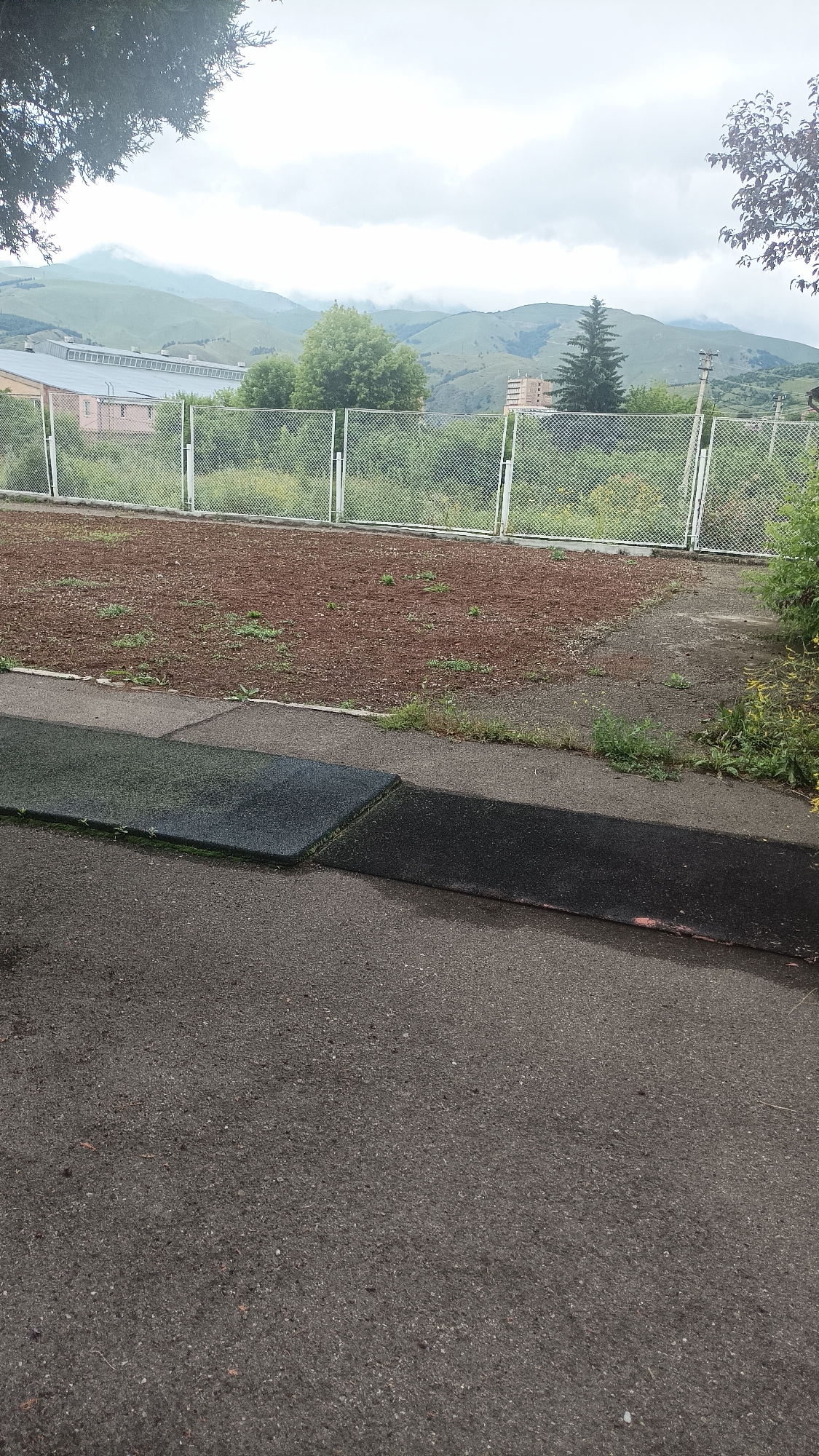 The image shows a view of the city of Vanadzor, Armenia, with a cloudy sky over rolling green hills and mountains in the background. In the foreground, there is a paved area with dark, rubberized surfaces. Beyond that, a chain-link fence encloses an area covered with reddish-brown soil and sparse vegetation. Further in the distance, behind the fence, a long, light-colored building with a dark roof is visible, alongside scattered trees and some taller structures. The overall scene depicts an outdoor environment, possibly a park or recreational area, with a backdrop of natural landscapes.