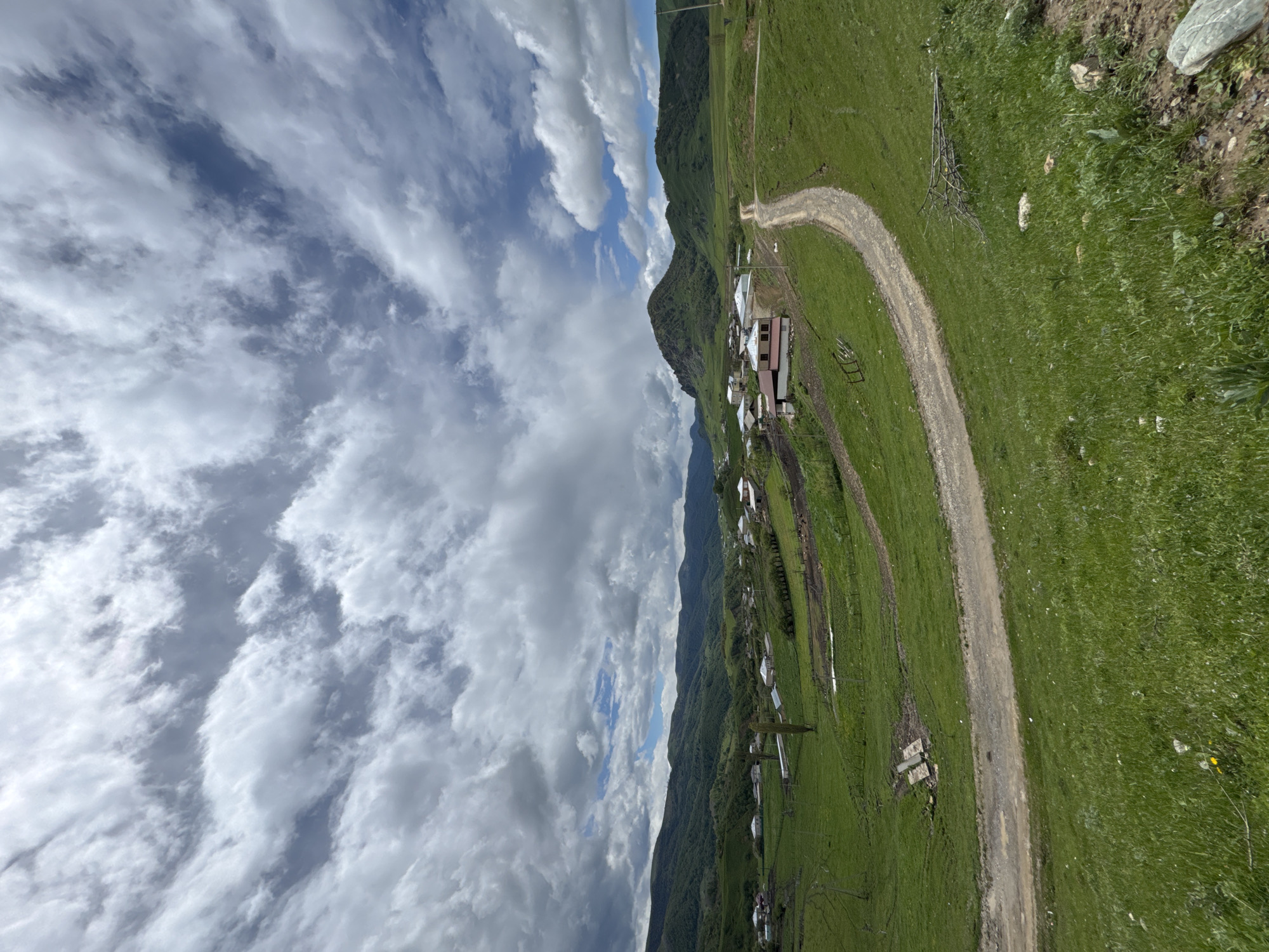 The image shows a rural scene in Верхняя Мара, Russia. A dirt road curves through a green grassy field, leading to a small village nestled along a mountainside. The village consists of several buildings with metal roofs. The sky is filled with clouds, partially obscuring the sky. The landscape consists of grass, soil, rocks, buildings, and mountains.