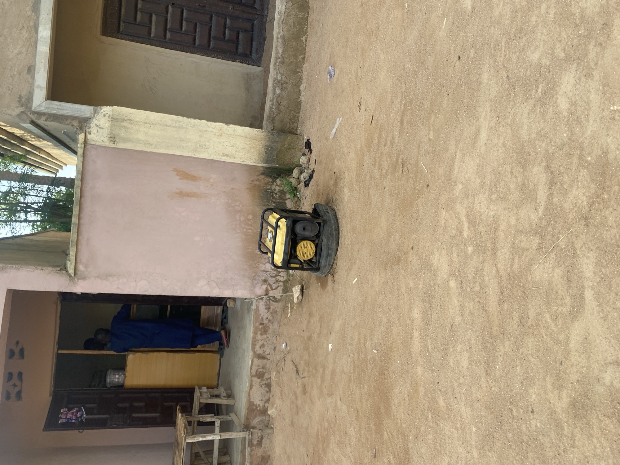 A generator sits on the ground in front of a building. The building has a pink-colored wall and a dark brown wooden door. To the left of the door, a person wearing a blue robe is seen inside the building. The ground is sandy and appears to be outdoors. There is some debris on the ground, including a few pieces of paper. The scene appears to be in a somewhat rural or less developed area. There is no visible text in the image. The lighting suggests it is daytime, and the weather appears to be clear.