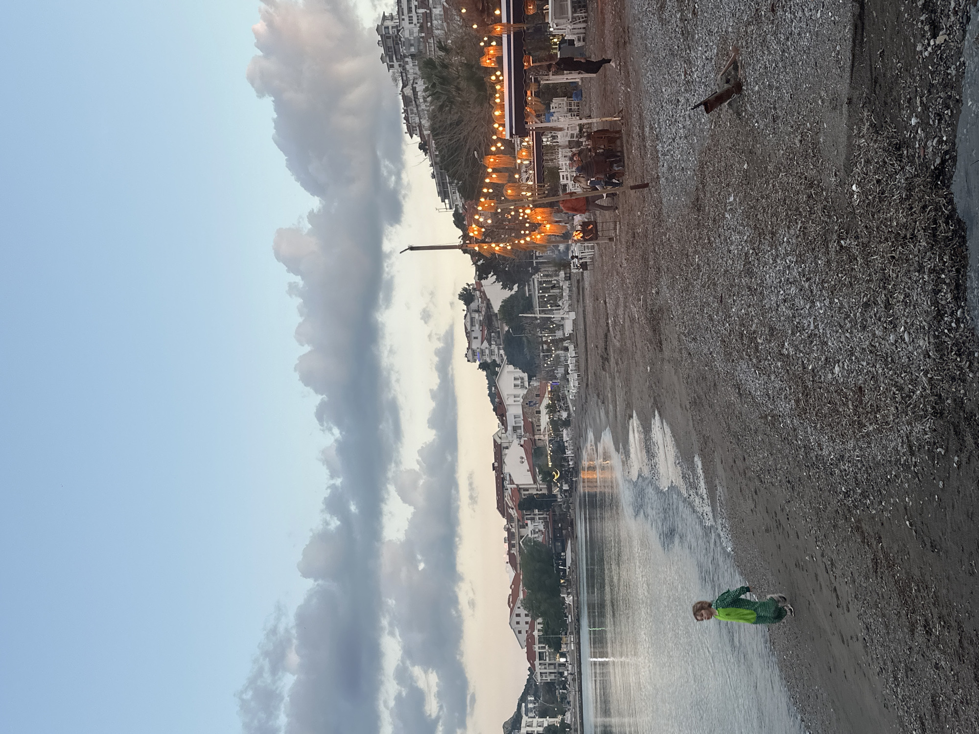 The image depicts a beach scene in Datça, Turkey, at dusk. A child in a green outfit is standing in the water near the shoreline. The beach is composed of dark sand or pebbles. Above the beach, there's a row of buildings that appear to be houses and restaurants. String lights illuminate an outdoor dining area of one of the restaurants. Above the buildings, the sky is filled with clouds, some tinged with the colors of sunset. In the background, more buildings are visible on higher ground, suggesting a hillside settlement. A long wooden pole stands erected in the foreground.
