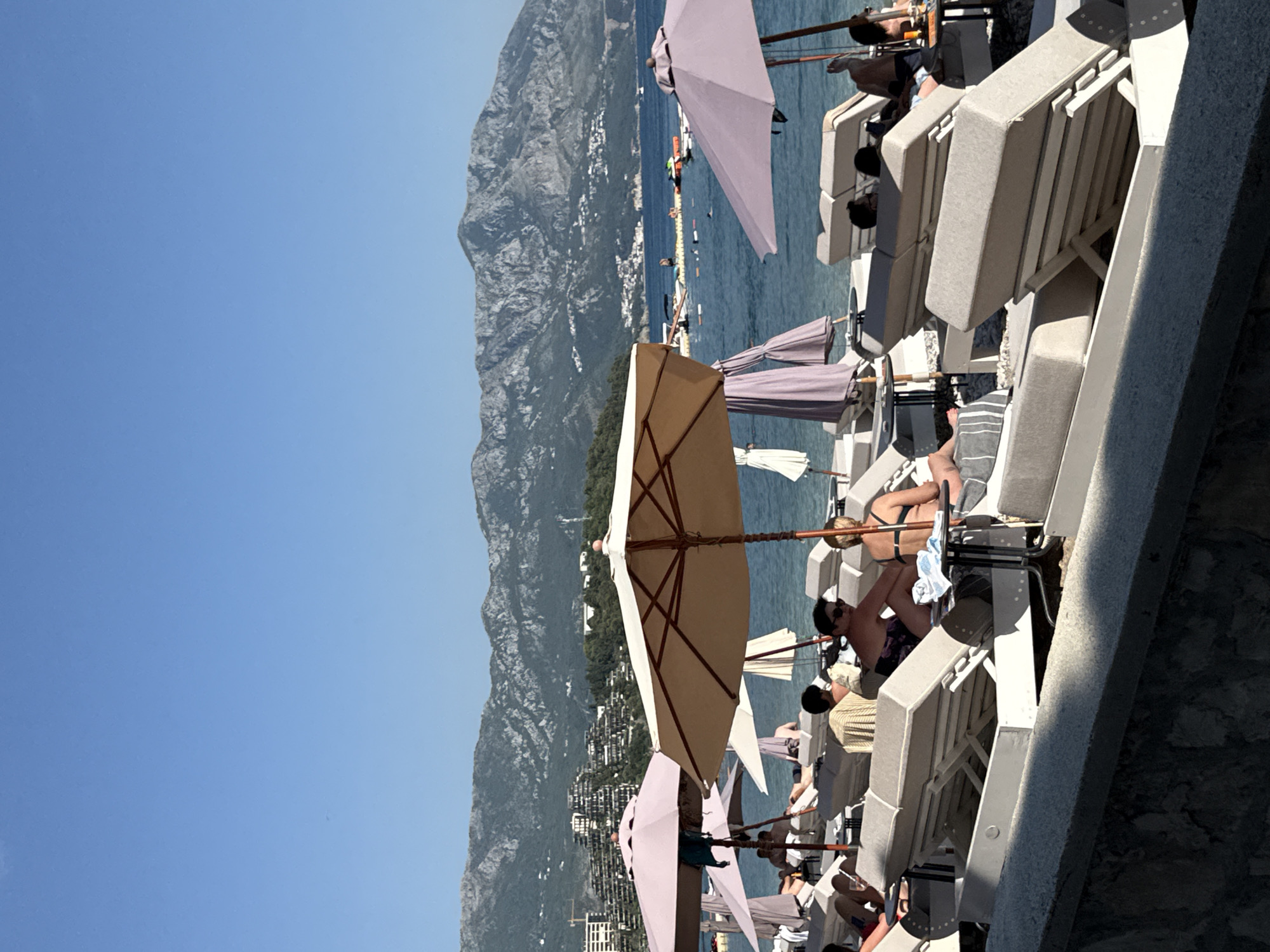 The image captures a sunny day at a beach in Budva, Montenegro, with a clear blue sky. In the foreground, several rows of empty and occupied lounge chairs are arranged on the pebbly shore, some shaded by large umbrellas. People are seen relaxing on the chairs, sunbathing, and some are engaged in conversation. A few small tables are placed between the chairs.
Beyond the beach, the calm, blue sea stretches out, dotted with several boats and a floating recreational area. In the background, a mountainous landscape rises, with buildings and structures visible on the slopes, indicative of the coastal city's development. The scene depicts a typical leisure scene at a popular tourist destination.