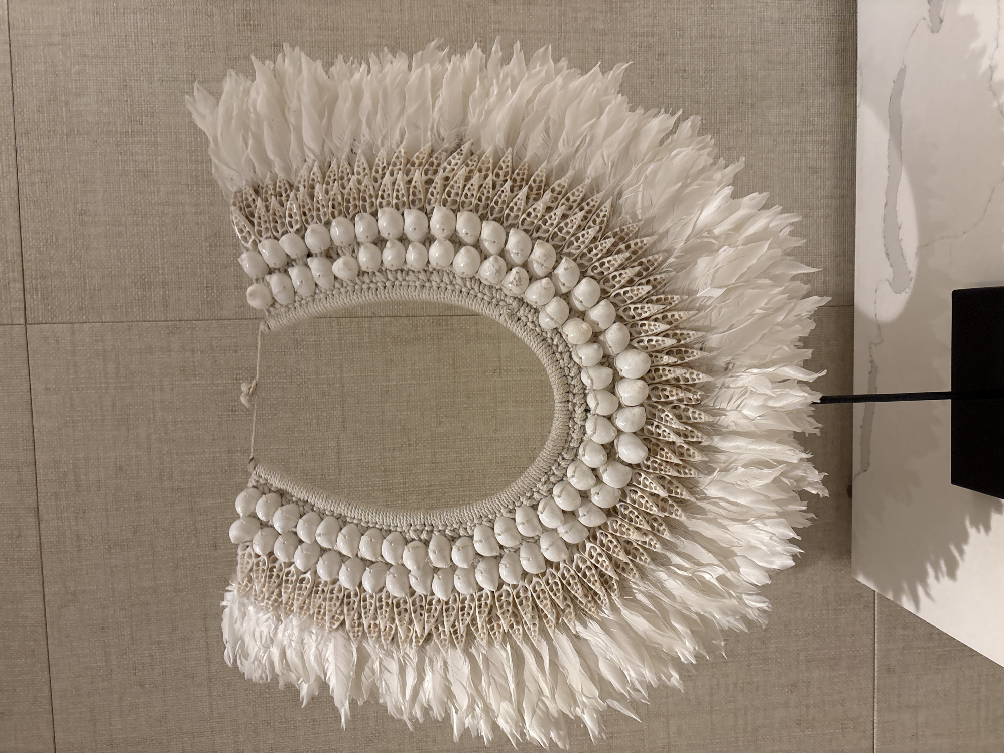 A close-up, eye-level, medium shot captures a decorative collar made of white feathers and shells, displayed against a textured, neutral-colored wall in Vagator, India. The collar, which resembles a neckpiece, is intricate, featuring rows of small, white, polished shells strung together and interspersed with delicately woven, tan-colored material. White feathers fan out from the outer edge, creating a voluminous, layered effect. The collar is supported by a thin, black metal stand, the base of which is partially visible on a white surface with subtle grey veining in the foreground, likely a countertop or table. The lighting is even, highlighting the textures and details of the collar. There are no people or animals present in the scene.