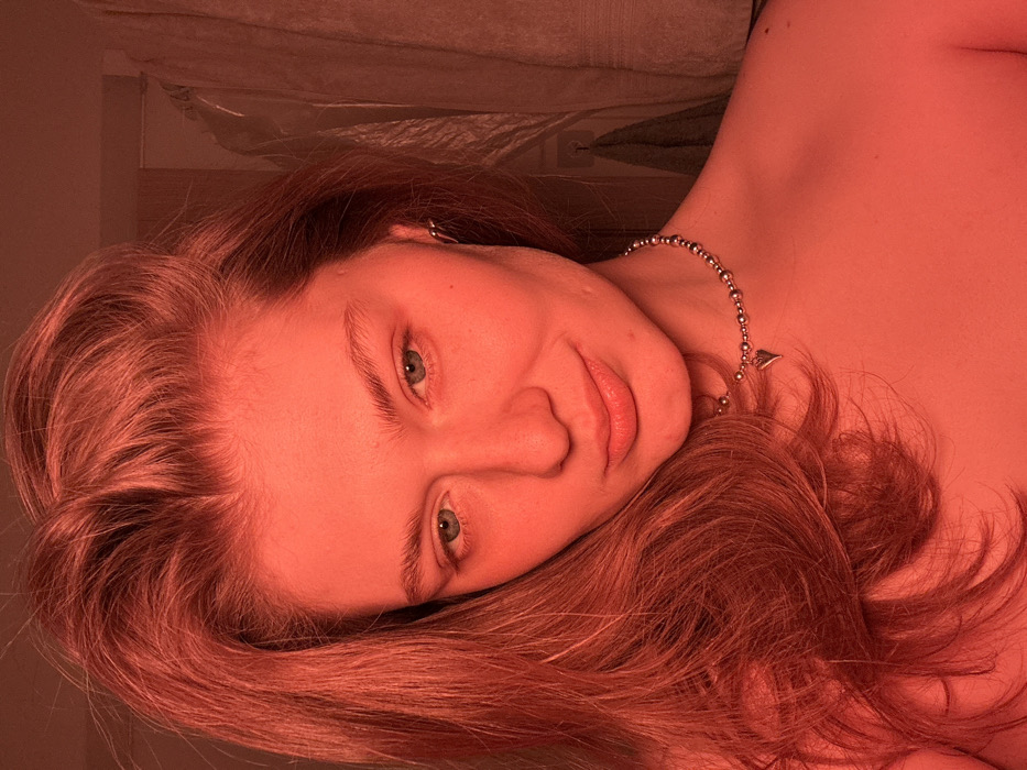 The scene, located in an indoor setting within the city of Noviy, Russia, captures a close-up portrait of a single female individual. The woman has long, light brown or reddish-brown hair, parted slightly off-center, cascading over her left shoulder and chest. Her face is directed towards the viewer, exhibiting light-colored eyes (possibly blue or green), natural eyebrows, and a slight smile. She wears a silver-colored beaded choker necklace with a small heart-shaped charm positioned on the lower right side of her neck. A small, silver-toned hoop earring is visible in her right ear. Her upper body, from the shoulders up, appears bare. The entire scene is heavily illuminated by a strong red light source, casting a pervasive monochromatic red hue over the subject and her immediate surroundings. The background is blurred and indistinct, revealing a lighter-toned textile hanging vertically on the far right, possibly a towel or garment, and a darker, textured surface on the left, possibly a wall or door frame. No other specific identifying objects or architectural details are discernible within the visible frame.
