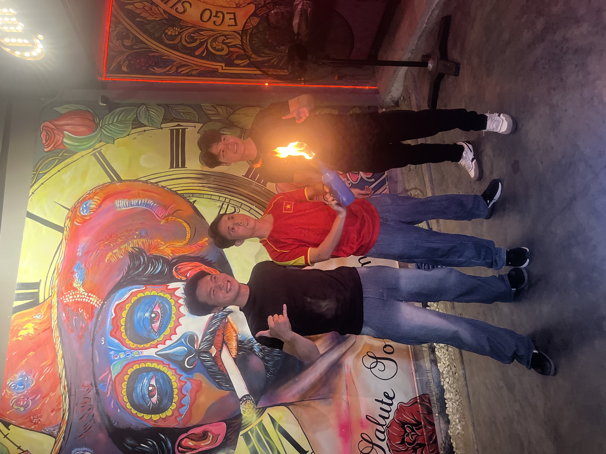 Three young Vietnamese men are posing in front of a colorful mural depicting a stylized sugar skull character with a large sombrero. The mural is vibrant, with bold colors and intricate details, including a clock face and a rose. The men are standing on a concrete floor. The man on the left is wearing a black t-shirt and gray pinstripe pants, giving a shaka sign with his left hand. The man in the middle is wearing a red t-shirt with the Vietnamese flag on it and blue jeans. He is holding a blue plastic bottle. The man on the right is wearing a black t-shirt and black pants, and he is holding a lit object that is emitting flames, which appears to be a fire-breathing trick. Behind the man on the right, a black industrial fan is visible. A neon sign with the word "EGO" in script and lights is also visible to the right of the mural. The overall impression is a casual gathering, possibly at a bar or entertainment venue in Đà Nẵng, Vietnam, given the cultural context suggested by the mural and the presence of a Vietnamese flag.