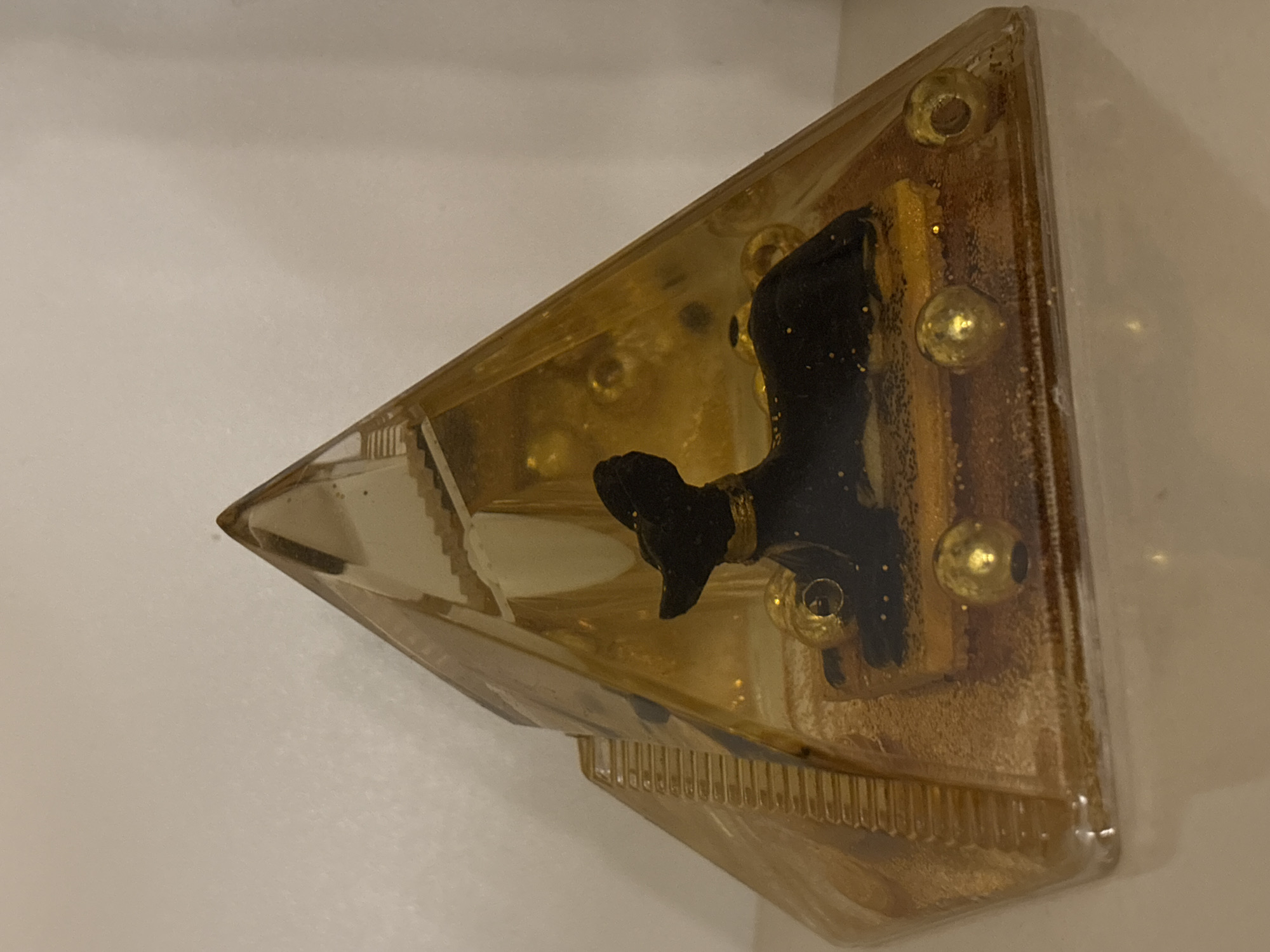 A triangular, golden-toned resin shelf is mounted on a white wall. Inside the resin are embedded decorative elements, including gold-colored spheres and a small, dark-colored dog figurine. A portion of a miniature staircase is also visible within the resin. The location is unspecified; the image provides no information about being in Sofia, Bulgaria.