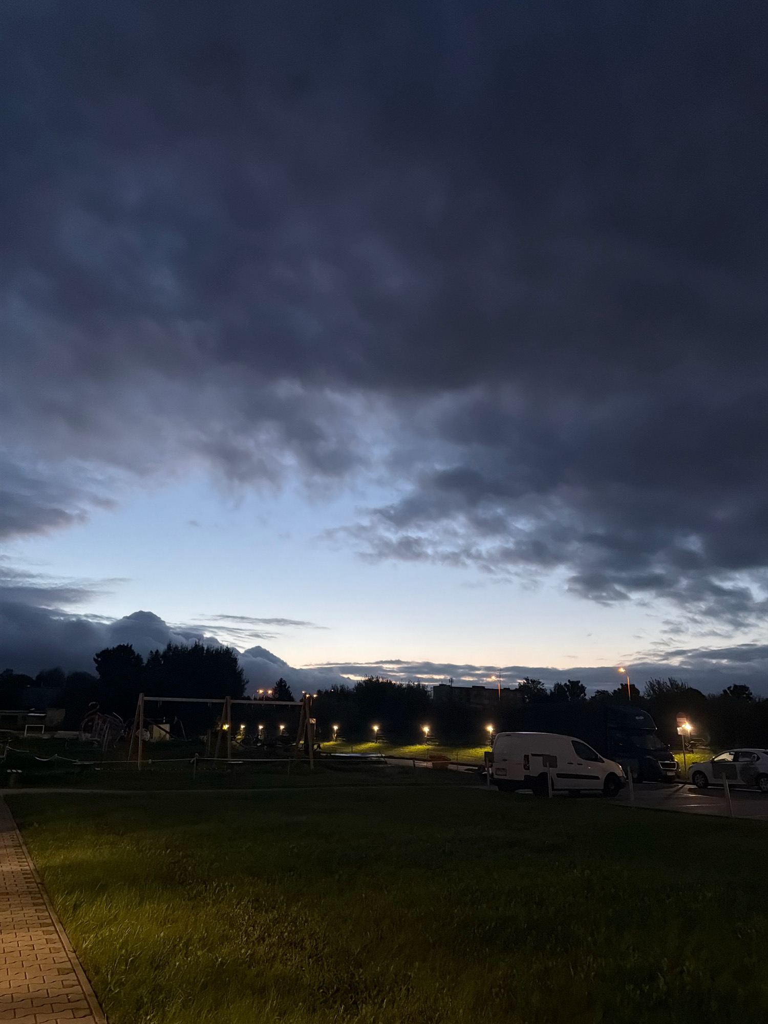 The image depicts a park area with a playground in the foreground. In the background, a van and a truck are parked. The sky is filled with dark, ominous clouds, suggesting an approaching storm. The overall atmosphere is dark and somber. The location is specified as Koszalin, Poland.