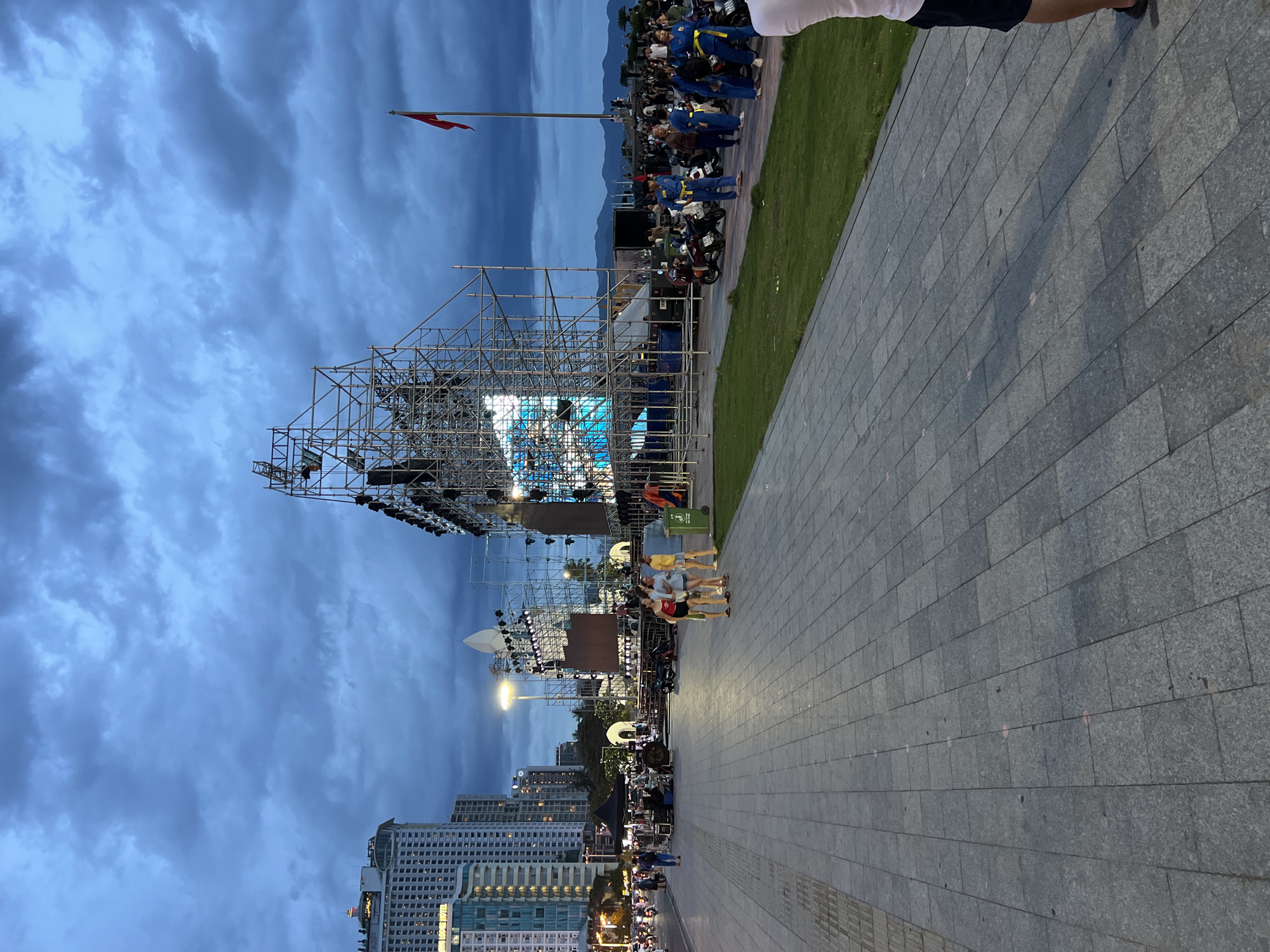The image depicts a broad, paved walkway in Nha Trang, Vietnam, bordering a green grassy area. In the background, tall buildings line the city's coastline. The main focus is a large, elaborate stage structure constructed from scaffolding, with a prominent screen displaying a bright blue image. A Vietnamese flag flies from a flagpole to the right of the stage. Several people are visible on the walkway and near the stage, including a group of women walking together, individuals in blue uniforms with yellow belts, and others milling about. Motorcycles are parked along the grassy area and on the walkway. The sky is overcast with dark, cloudy conditions, suggesting either dusk or an impending storm. The scene appears to be set up for an outdoor event or performance.