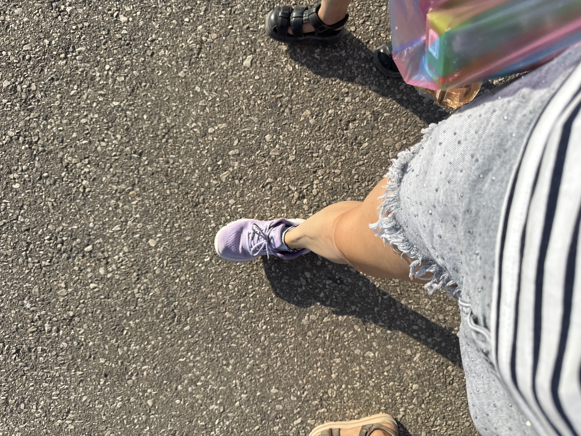A first-person perspective shot captures a person's legs and feet walking on an asphalt surface. The person is wearing a grey denim skirt with frayed edges, adorned with small sparkling studs. A striped t-shirt is visible at the bottom of the frame. On the right leg, a purple sneaker with white laces is worn. To the left of the frame, another person's leg is partially visible, wearing a sandal. To the right, another person is wearing black chunky sandals. The scene is lit by bright sunlight, casting shadows on the ground. The environment appears to be an outdoor urban setting, possibly a street or walkway in Yevpatoriya, Ukraine.