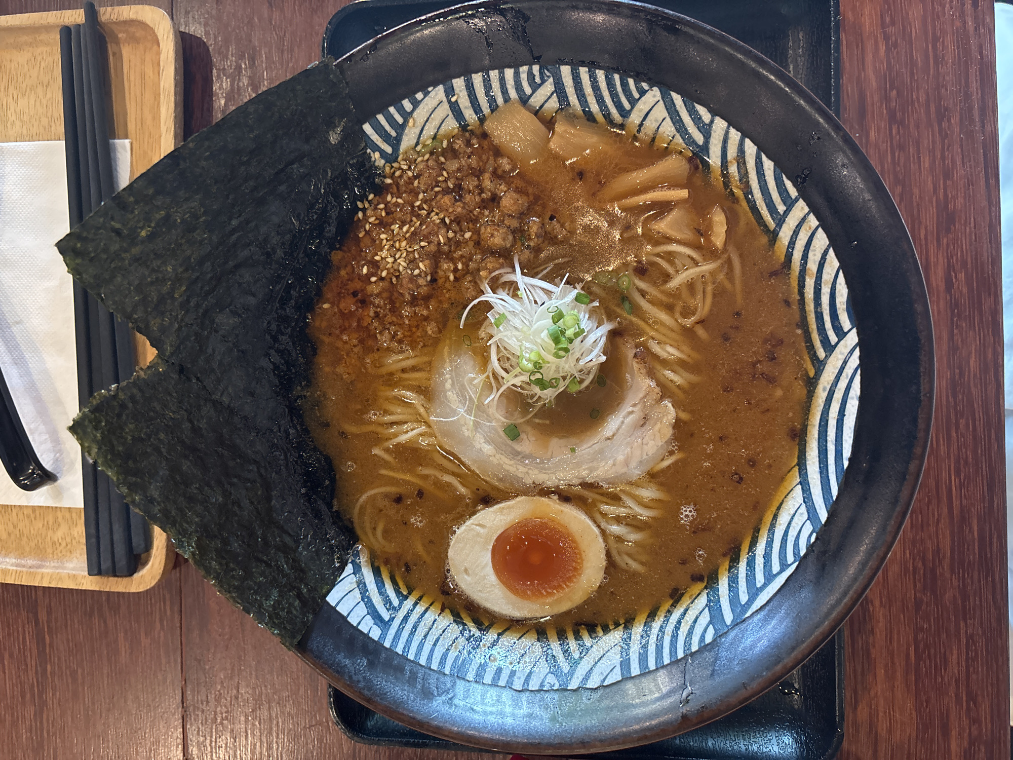 An overhead shot depicts a bowl of ramen on a wooden table in Phuket City Municipality, Thailand. The ramen bowl, featuring a decorative blue and white wave pattern around the rim, contains noodles, broth, half a soft-boiled egg, a slice of pork, bamboo shoots, ground meat sprinkled with sesame seeds, and shredded green onions. To the left of the bowl, two pieces of dried seaweed are positioned, with a small wooden tray holding a pair of chopsticks and a white napkin. The table is made of dark brown wood. The bowl rests on a black rectangular tray.