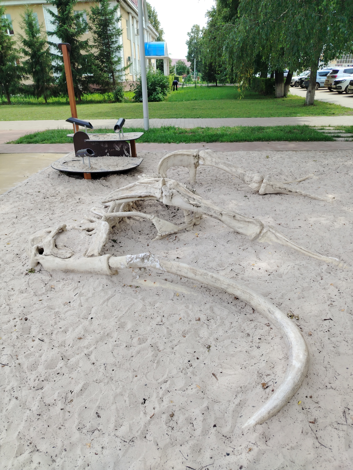 In Yanaul, Russia, a replica of a dinosaur skeleton is partially buried in a sandy playground area. The skeleton, consisting of a large skull, rib cage, and limbs, is made of a light-colored material, possibly concrete or fiberglass. A wooden pole with black tubes is positioned behind the skeleton, part of a playground apparatus. The playground is surrounded by a grassy area with trees and a building in the background. A person in dark clothing is visible in the distance, near the building. The ground is covered in sand, with scattered debris and footprints. The sky is overcast.