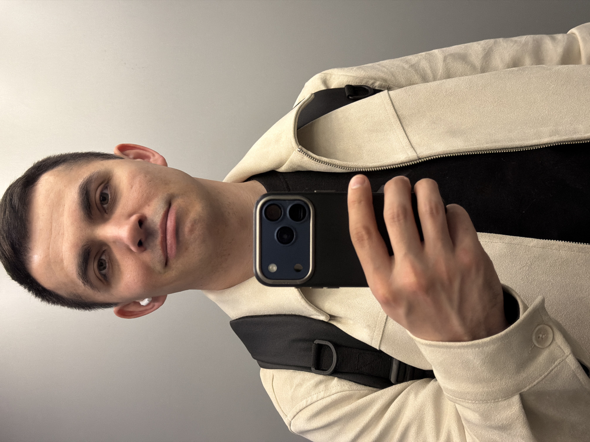 A man is taking a selfie. He is wearing a black shirt, a beige jacket, and a black backpack. He is holding a black smartphone with three camera lenses. The background is a plain, light-colored wall. There is no visible information to indicate this image was taken in Izhevsk, Russia.