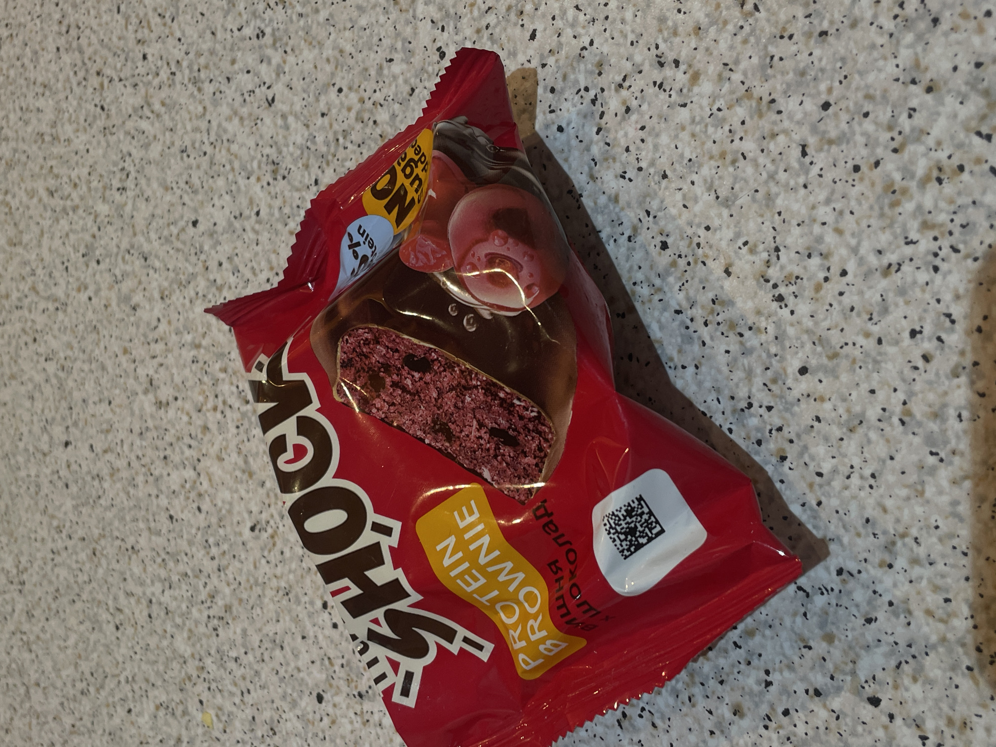 The image displays a red packaged food item, likely a snack bar or brownie, resting diagonally on a light speckled countertop. The background is a textured surface, possibly granite or a similar material, with a mix of light gray, white, and black specks.
The primary subject is the red wrapper, which features a prominent graphic of the product itself: a cross-section of a dark reddish-purple brownie-like interior coated in chocolate, adorned with what appears to be cherries or a cherry-flavored topping.
Visible text on the wrapper includes the brand name "Shook" in large white letters with a brown outline, positioned on the left. A yellow banner across the bottom right reads "PROTEIN BROWNIE". In the upper left corner, a blue circular label states "NO SUGAR ADDED", though part of it is obscured. A small white square containing a black QR code is visible on the right side of the wrapper. Below the QR code, some Cyrillic text is partially visible, which includes characters that suggest words like "шоколад" (chocolate) and "вишня" (cherry), aligning with the product's visual description.
The setting appears to be indoors, possibly a kitchen or dining area, indicated by the countertop. There is no specific activity taking place; it's a static image of the packaged item. Lighting is bright and seems to come from an overhead source, casting a slight shadow behind and to the right of the wrapper. The wrapper is slightly crinkled, indicating it's a soft, flexible material. The overall impression is a clear, well-lit shot of a product designed for a healthy-conscious consumer, given the "PROTEIN" and "NO SUGAR ADDED" claims.