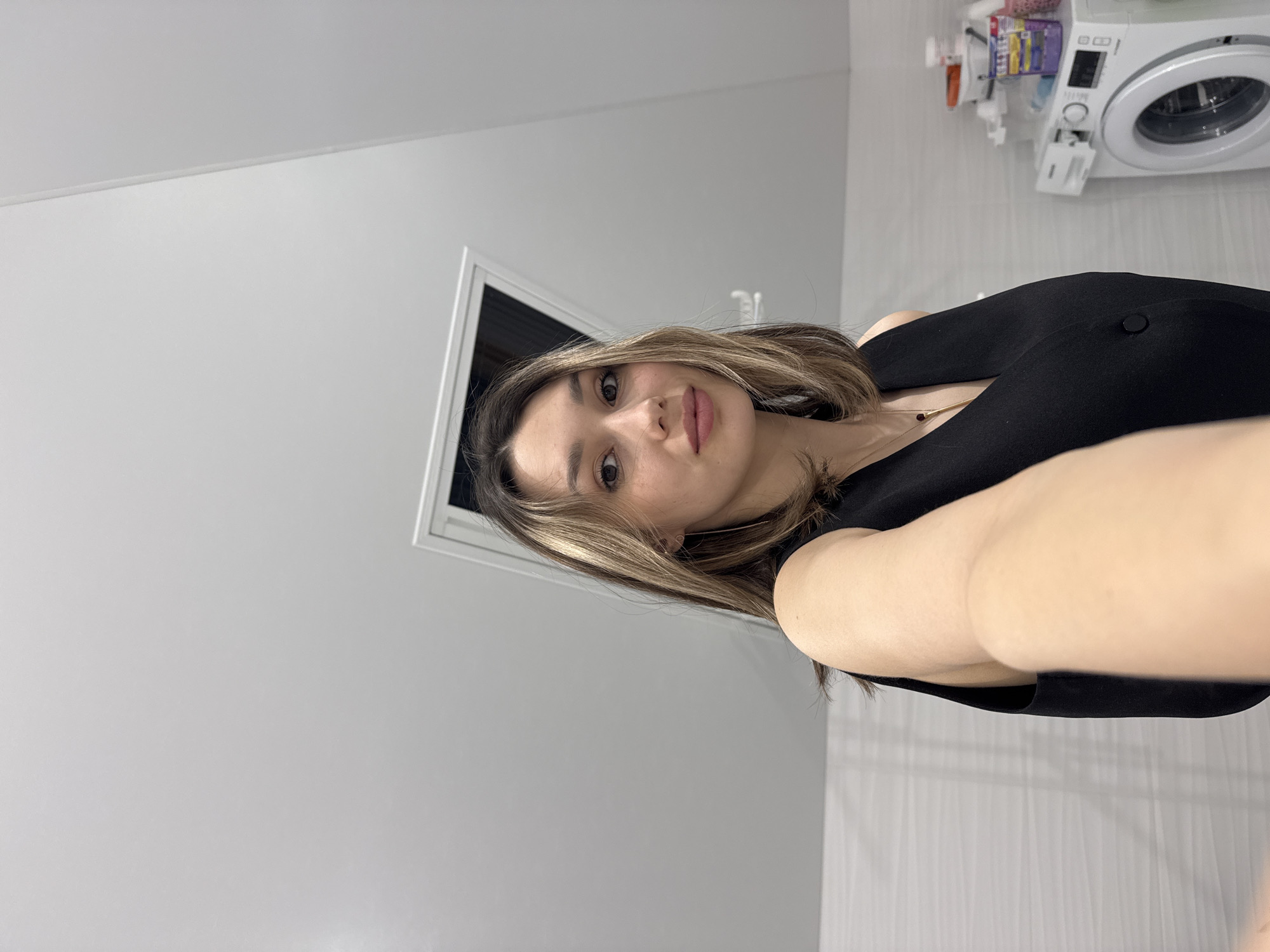 A medium, eye-level shot shows a young woman holding a camera at arm's length, presumably in a self-portrait. The location appears to be a bathroom. The walls are light grey, and a small, framed window is visible behind her head. A white washing machine and various cleaning supplies can be seen in the upper right corner of the image. The subject has fair skin, brown hair with blonde highlights, and is wearing a black top. A delicate gold necklace is also visible. The exact city location is unknown.