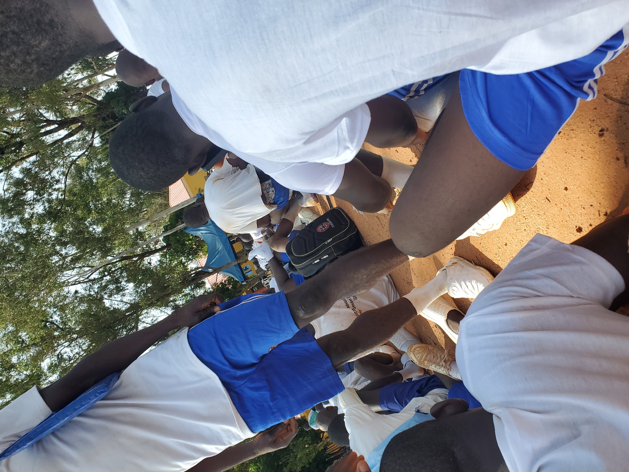 In Barakin Rafin Gora, Nigeria, a group of young people, likely students, are gathered outdoors on a dirt surface, possibly a schoolyard. The scene is partially obscured by the perspective, focusing on the lower bodies and the ground. The individuals are wearing white t-shirts and blue shorts, some with white stripes. Several are sitting or kneeling, with some visible wearing white socks and sneakers. A black backpack with a white logo is on the ground near some of the individuals. The background shows trees with lush green foliage and a glimpse of a red-roofed building and a blue tent. The lighting suggests it is daytime, with sunlight casting shadows on the ground.