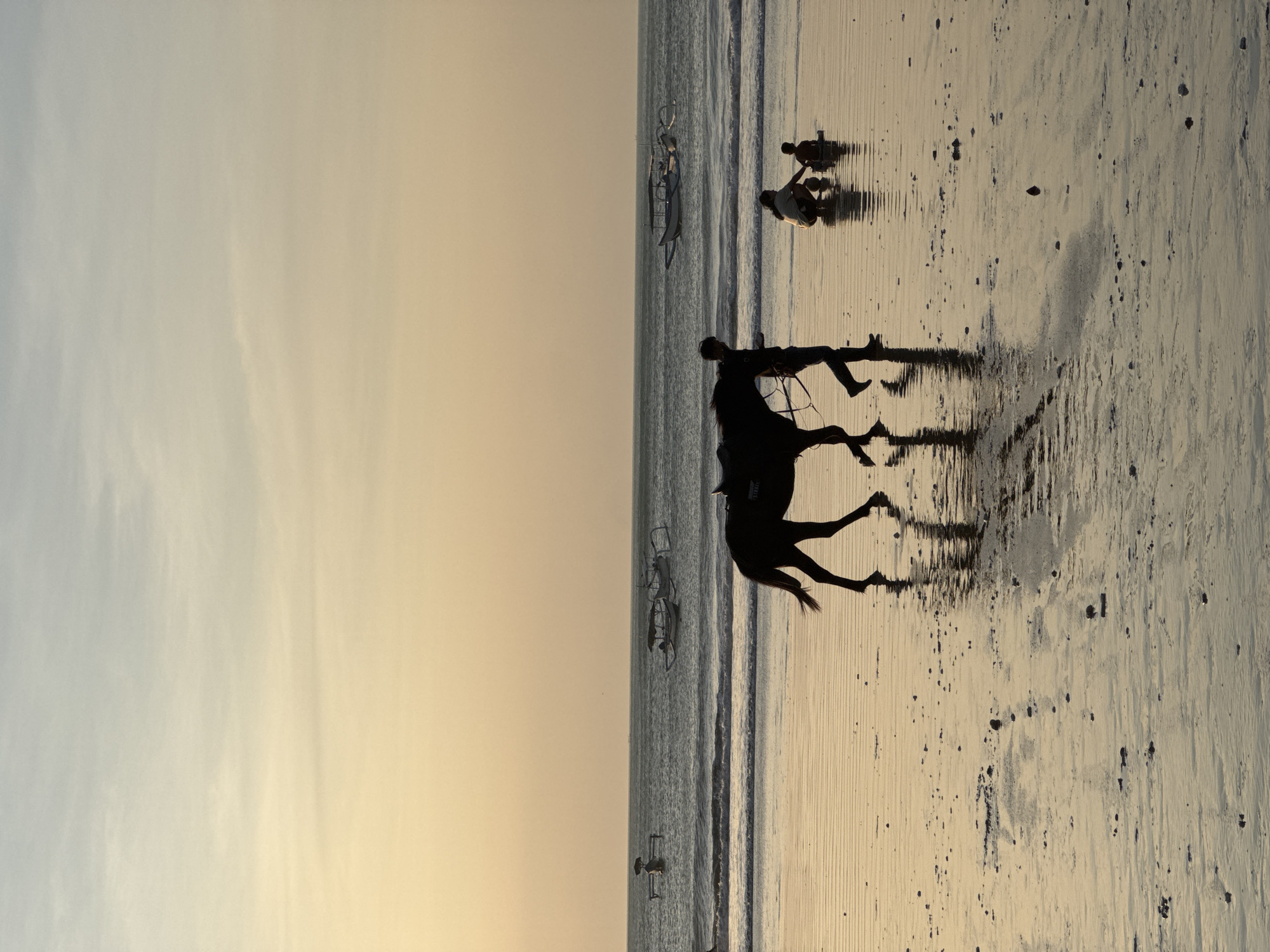 A silhouette of a person leading a horse walks along the wet sand of a beach at sunset. The horse is dark and appears to be wearing a saddle. The person is walking to the right of the horse. In the background, the ocean is calm with a few boats anchored offshore. Two people are crouched on the sand to the right of the horse, appearing to interact with each other. The sky is a soft gradient of yellow and pale blue, typical of a sunset in Kuta Selatan, Indonesia. The wet sand reflects the light and the figures, creating a serene and atmospheric scene.