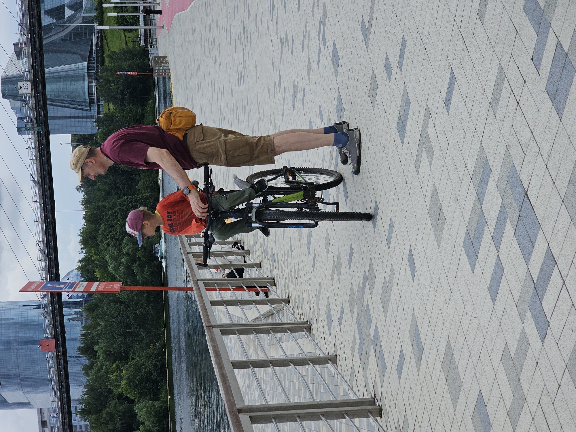 A man and a boy are standing on a paved walkway near a body of water. The man, wearing a maroon t-shirt, tan shorts, and a tan cap, has a yellow fanny pack around his waist. He is looking to his left. The boy, wearing a red shirt with "COOL BOY" printed on it and a purple and white baseball cap, is sitting on a bicycle. The bicycle is positioned in front of the man, and the boy's hands are on the handlebars. The walkway is paved with geometric patterns of light and dark gray tiles. A metal railing runs along the edge of the walkway, separating it from the water. In the background, there are lush green trees, modern buildings with glass facades, and a bridge. The sky is overcast. The scene appears to be a public area in the city of Khimki, Russia, likely along the Moscow Canal, given the architectural style and the presence of modern buildings and a bridge.