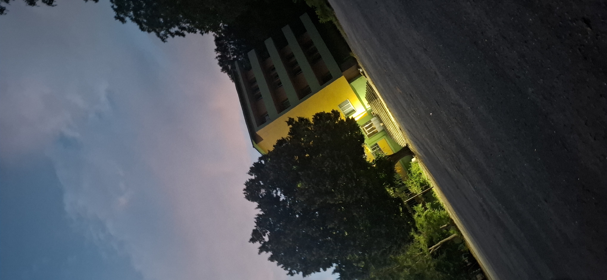 The image shows a street in the city of Shumen, Bulgaria, with a building and a large tree. The building is tall and has a yellow facade with green balconies and window frames. The tree is dark and leafy, positioned in front of the building. The sky is dusky with hues of blue and pink, suggesting it is either dawn or dusk. The street in the foreground is asphalt and appears to be angled towards the bottom right of the frame. The lighting suggests artificial lights are on in the building, illuminating the entrance and some of the lower floors. There are no people visible in the image.