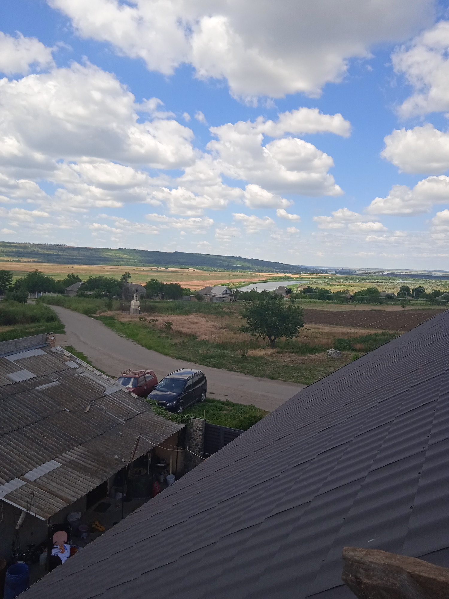 The image depicts a rural landscape under a bright, cloudy sky. In the foreground, two rooftops are visible; the one on the left is made of weathered, corrugated sheeting, while the one on the right is a darker, more modern material. Adjacent to the left rooftop, a small, open-air structure or shed contains various items, including buckets, a chair with clothing draped over it, and a blue barrel. A paved road leads away from the foreground, curving into the middle ground where two parked cars, a red hatchback and a dark SUV, are situated.
Beyond the road, the scene opens up to a sprawling vista of fields, trees, and scattered houses, characteristic of a village. The fields appear to be a mix of cultivated land and dry grass, with patches of darker soil suggesting recent plowing. A line of trees marks the edge of the cultivated area, and further in the distance, a river or large body of water can be seen, reflecting the sky. The horizon is defined by rolling hills covered in vegetation. The architecture of the houses suggests a traditional rural style. The overall impression is one of a peaceful, sunlit day in a Ukrainian countryside.