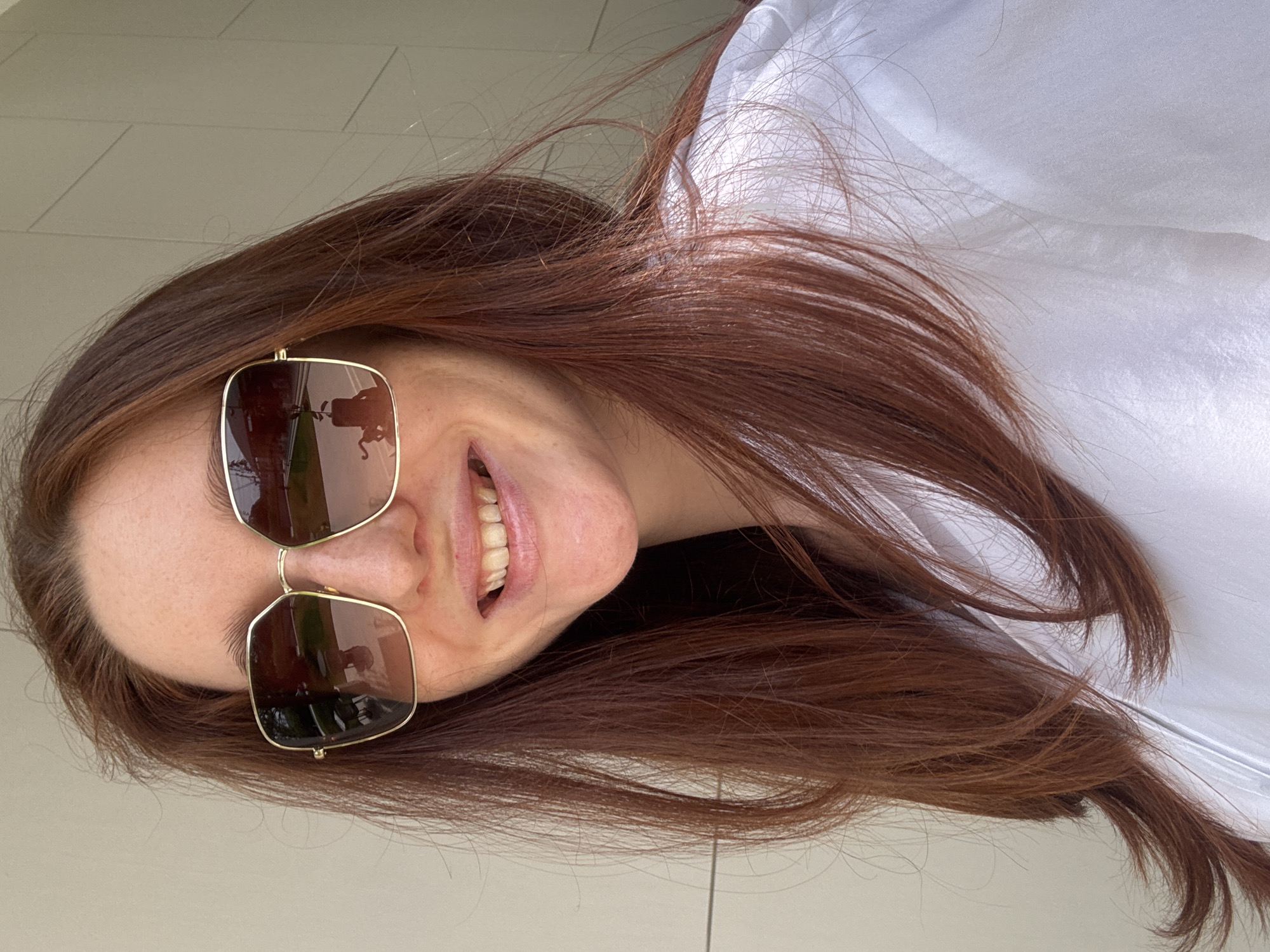 A woman with auburn hair, wearing brown square sunglasses and a white shirt, smiles at the camera. The reflection in her sunglasses suggests she is outdoors, possibly in Choeng Thale, Thailand, given the tile floor and architectural style that might be consistent with the region.