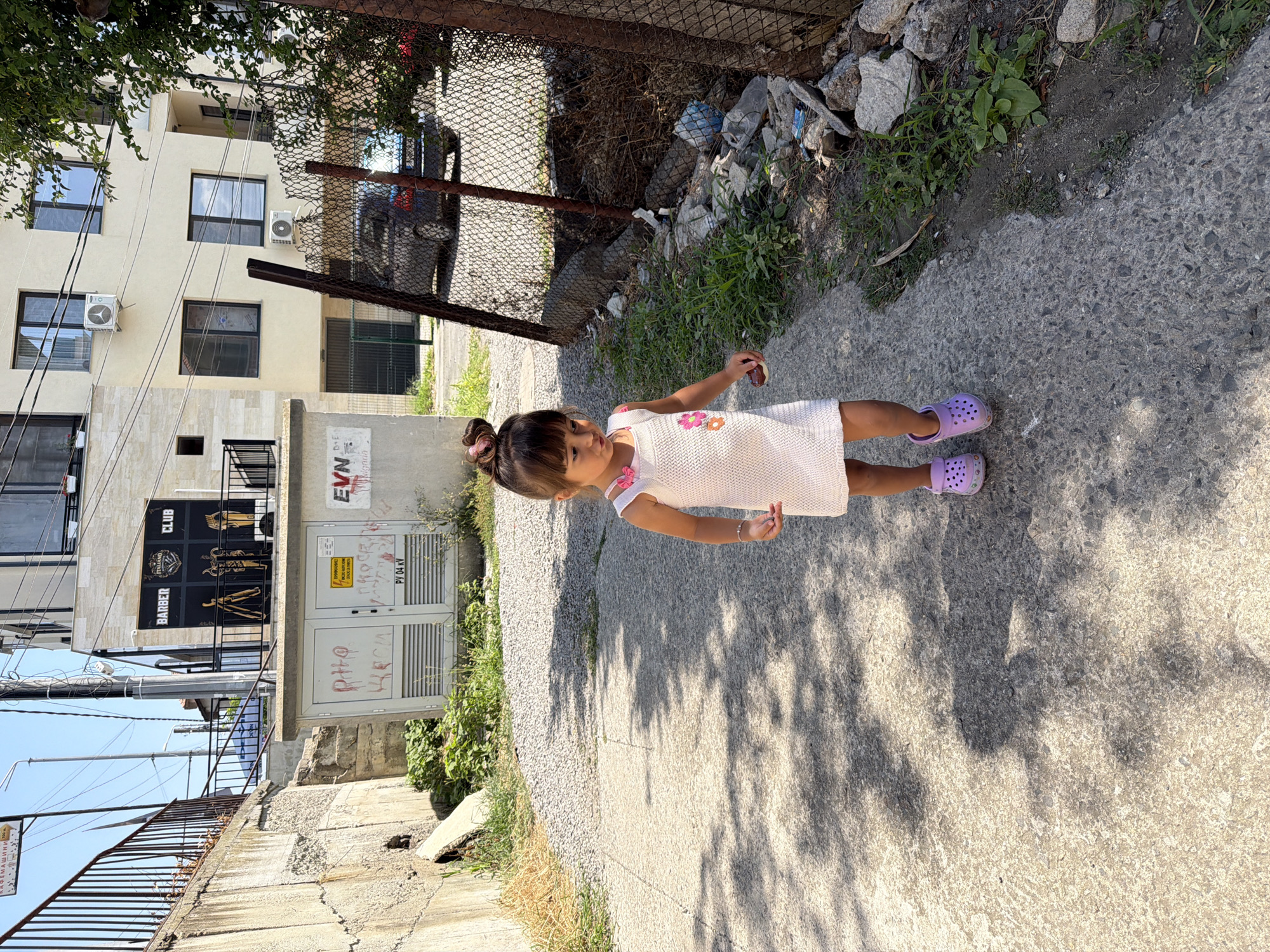 A young girl with dark hair tied in a bun, wearing a pink and white dress and purple Crocs, walks along a concrete path in Burgas, Bulgaria. She is looking down and to her left, holding a small, dark object in her right hand. To her left, a concrete wall with overgrown vegetation rises, and beyond that, a building with a sign for "BARBER CLUB ITALIANO" is visible. To her right, a chain-link fence separates the path from an area with debris, including plastic bottles, rocks, and dry branches. A dark-colored car is parked behind the fence. The scene is illuminated by sunlight, casting shadows on the path.
