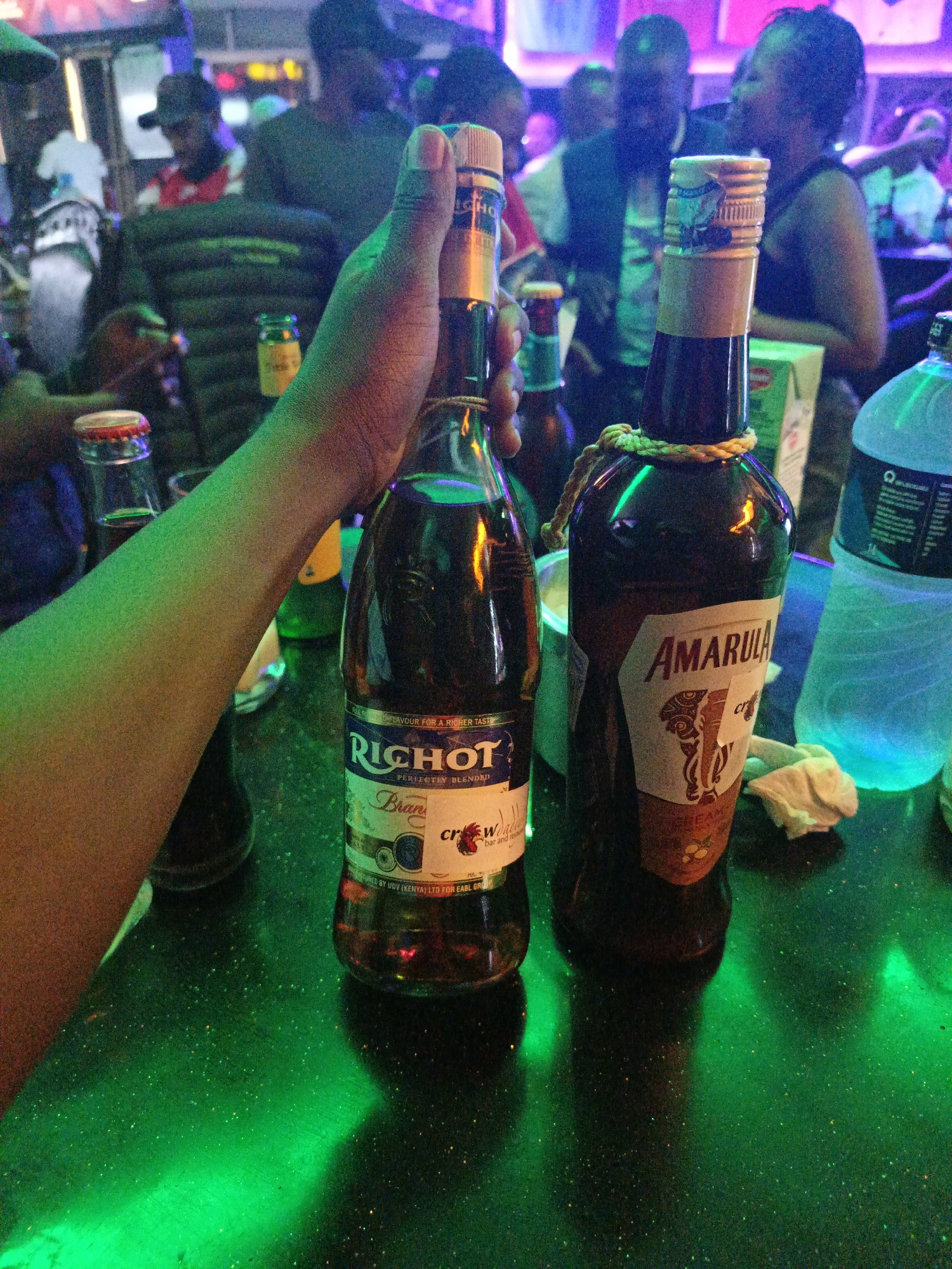The image depicts an indoor scene within a nightlife establishment in Nairobi, Kenya. The exact street or establishment name is not visually discernible. In the foreground, a dark-skinned arm is holding a clear glass bottle of Richot Brandy. To the right, a dark glass bottle of Amarula Cream liqueur, adorned with an elephant logo and a decorative rope around its neck, stands on a dark, reflective countertop. The Richot Brandy bottle's label indicates it is manufactured by "UDV (KENYA) LTD." The countertop is speckled and brightly illuminated by green light. Partially visible objects on the counter include a small bottle with a red and silver cap, a green bottle behind the arm, a crumpled white tissue, and a large translucent blue plastic bottle, likely containing water or a mixer, on the far right. The blurred background features multiple individuals of varying complexions, casually dressed, indicating a social gathering. The environment is characterized by mixed purple, blue, and green neon lighting, consistent with a bar or club setting.