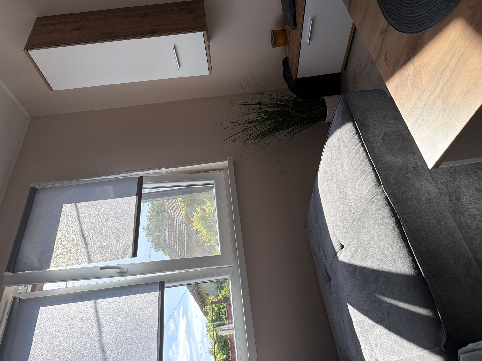 The image depicts an interior scene of a room. A window with grey roller blinds is visible, offering a view of greenery outside, possibly trees. A grey sofa is positioned on the right side of the image, with a potted plant behind it near the window. Overhead are two cabinets, one with a wood-grain finish and a white door, and another adjacent to it with a combination of white and wood-grain finishes. The lighting is a mix of natural light coming through the window and some artificial lighting. The room appears to be in Litakovo, Bulgaria, as per the request, but there is no intrinsic evidence in the image to confirm this location specifically.