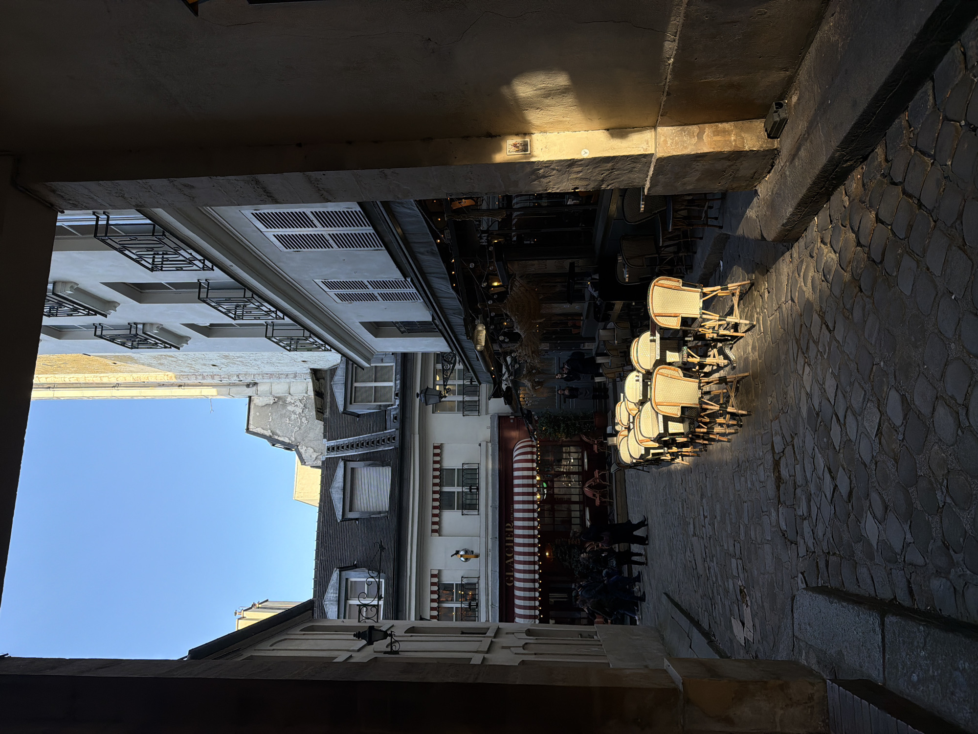 The scene depicts a narrow, cobbled street or alleyway in Paris, France, viewed from within a shadowed architectural passage. Strong, low-angle sunlight illuminates the right side of the street and buildings, casting long shadows and leaving the immediate foreground and left side in deep shade.
The street is paved with irregularly shaped cobblestones. Along the right side, a stone curb borders an outdoor seating area. This area features multiple empty tables and chairs with light-colored woven backs and dark frames, arranged in rows. Above these, a dark awning is visible, decorated with string lights and hanging straw-like elements. The adjacent building facade on the right exhibits multiple stories with white louvered shutters and black wrought-iron balconies. A small, framed picture is affixed to a sunlit pillar on this facade. A dark rectangular object rests on the curb in the lower right.
In the mid-ground, a white building displays a red and white striped awning over a storefront partially labeled "GLACIER." A gold-colored, duck-like ornament is mounted on the wall above the awning. This building features windows with small balconies on its upper floors and dormer windows on its roof.
A group of approximately five to seven individuals, dressed in dark attire, are walking away from the viewer on the cobblestone street, primarily concentrated in the mid-ground near the "GLACIER" establishment. Other figures are indistinctly visible further down the street or near the entrance of the right-side establishment.
On the left, a portion of a building with a light-colored facade and a black, ornate streetlamp is visible. The upper sections of buildings frame a clear, bright blue sky.
