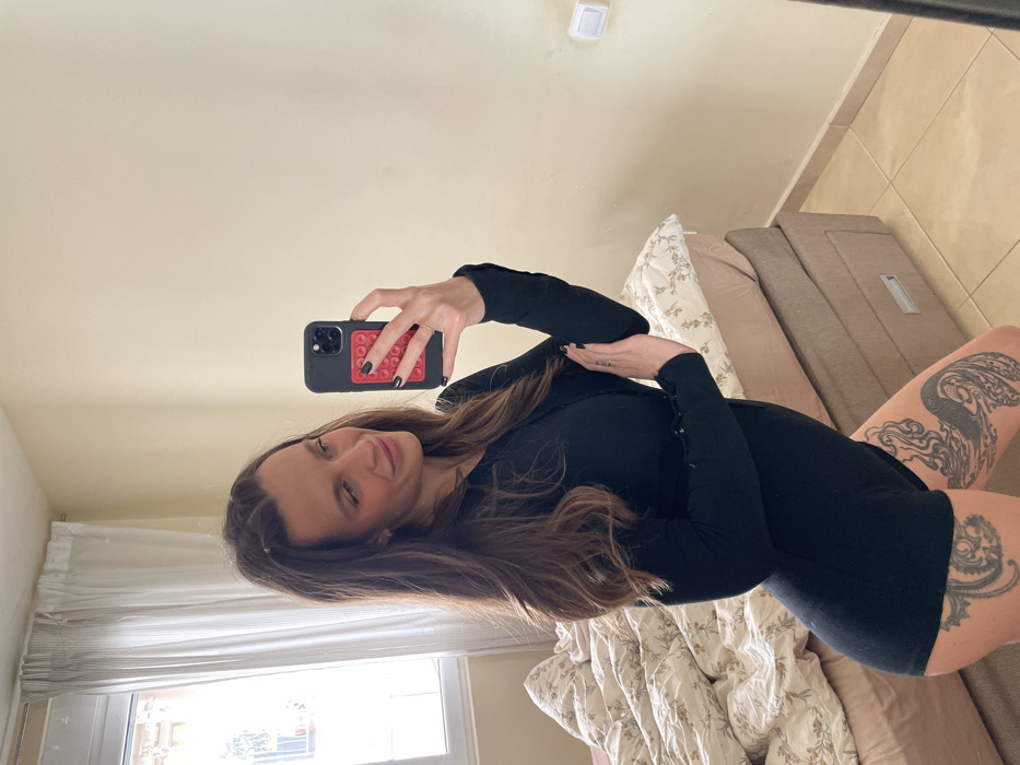 The image depicts an adult female subject taking a mirror selfie within an indoor setting, identified as a room in the city of Budva, Montenegro.
The subject is a light-skinned female with long, dark brown hair. She is wearing a tight-fitting, long-sleeved black romper or bodysuit. Visible on her upper thighs are extensive, intricate dark-inked tattoos, appearing as a large, flowing design on her left thigh and a similar detailed pattern on her right thigh. Her fingernails are painted black, and she wears a ring on the ring finger of her left hand. She holds a dark-colored smartphone with her right hand, which features a red, square-shaped pop-it style accessory on its back. Her left arm is bent, with her left hand resting on her right bicep. She gazes towards her reflection in the mirror, exhibiting a slight smile.
The background shows a bedroom environment. To the subject's left is a bed with a light brown frame, including two storage drawers visible at its base. The bed is covered with a patterned duvet and pillows, featuring a light background with subtle floral or leafy designs. The bedding appears somewhat disheveled. The walls are painted a cream or light beige color. In the upper left background, a window is visible, covered by white translucent curtains, through which natural light enters and a blurred view of an exterior building structure can be discerned. To the subject's right, on the cream wall, a white electrical light switch is present. The floor is composed of light-colored, square tiled sections. The lighting suggests a daytime setting.