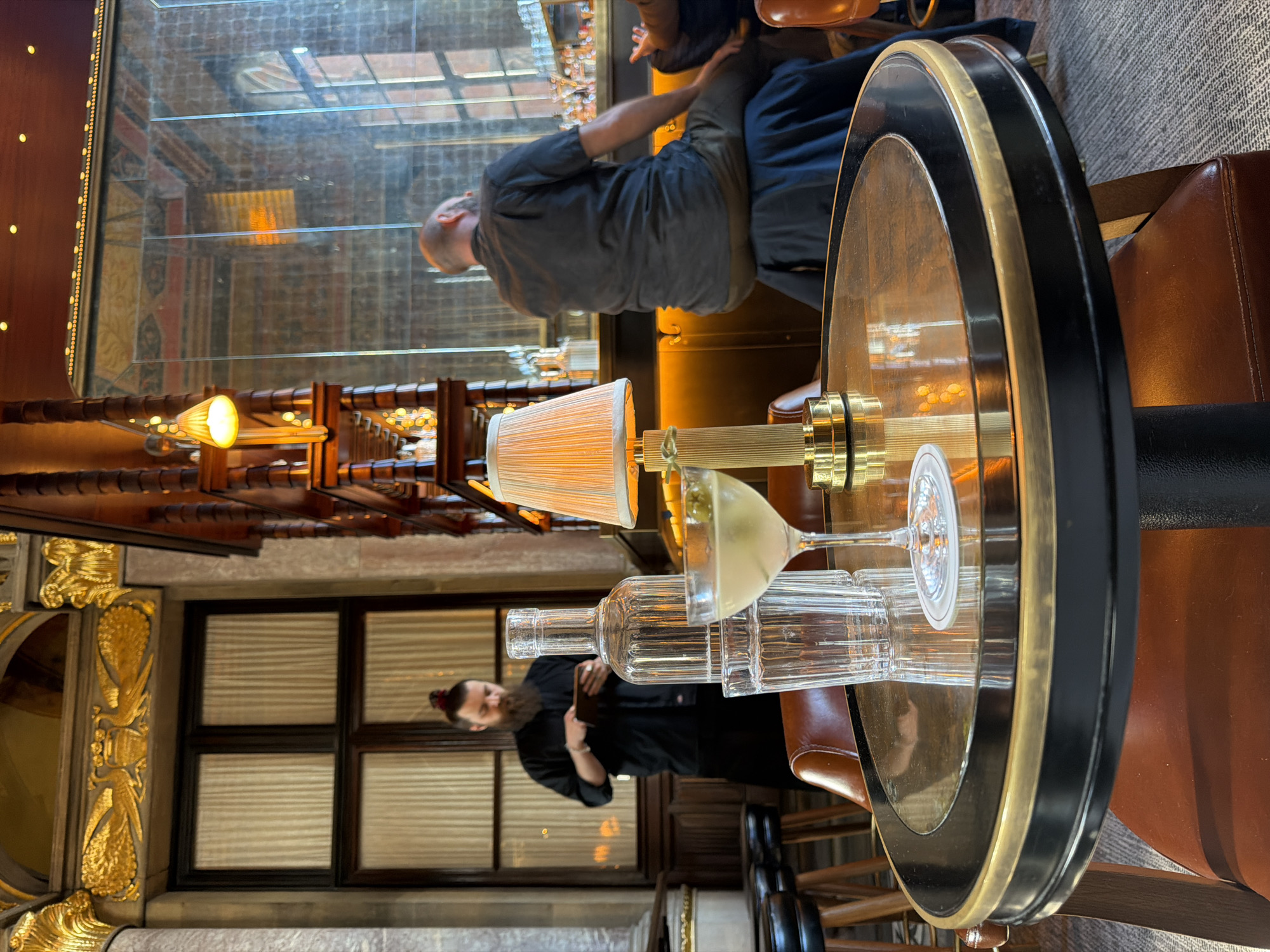 This image captures an indoor scene within an upscale bar or restaurant in London, United Kingdom, likely during the evening. The setting is characterized by warm lighting and rich textures, creating a sophisticated ambiance.
In the foreground, a polished round table features a dark, possibly wooden, top with prominent gold and black circular rims. On the table, a small lamp with a pleated, light-colored shade emits a soft glow. Adjacent to the lamp, a martini glass filled with a pale yellow drink and garnished with an olive is displayed, alongside a tall, clear glass bottle and two empty, fluted glasses. A portion of a brown leather chair is visible next to the table.
To the left, a man with a dark beard and hair tied in a bun, wearing a dark jacket or shirt, stands and looks down at something held in his hands, possibly a menu or a mobile device. Further into the background, another man with a bald head and a beard, dressed in a dark top, is seated at a bar counter, facing away from the camera. The bar area is softly lit, with various bottles and glassware visible behind the counter.
Architectural details include ornate golden carvings on a wall section to the far left, and dark-framed windows with what appear to be blinds or frosted glass panels. Wooden, lattice-like structures adorned with small lights are also part of the interior design, enhancing the warm and inviting atmosphere. No discernible text is visible in the image.