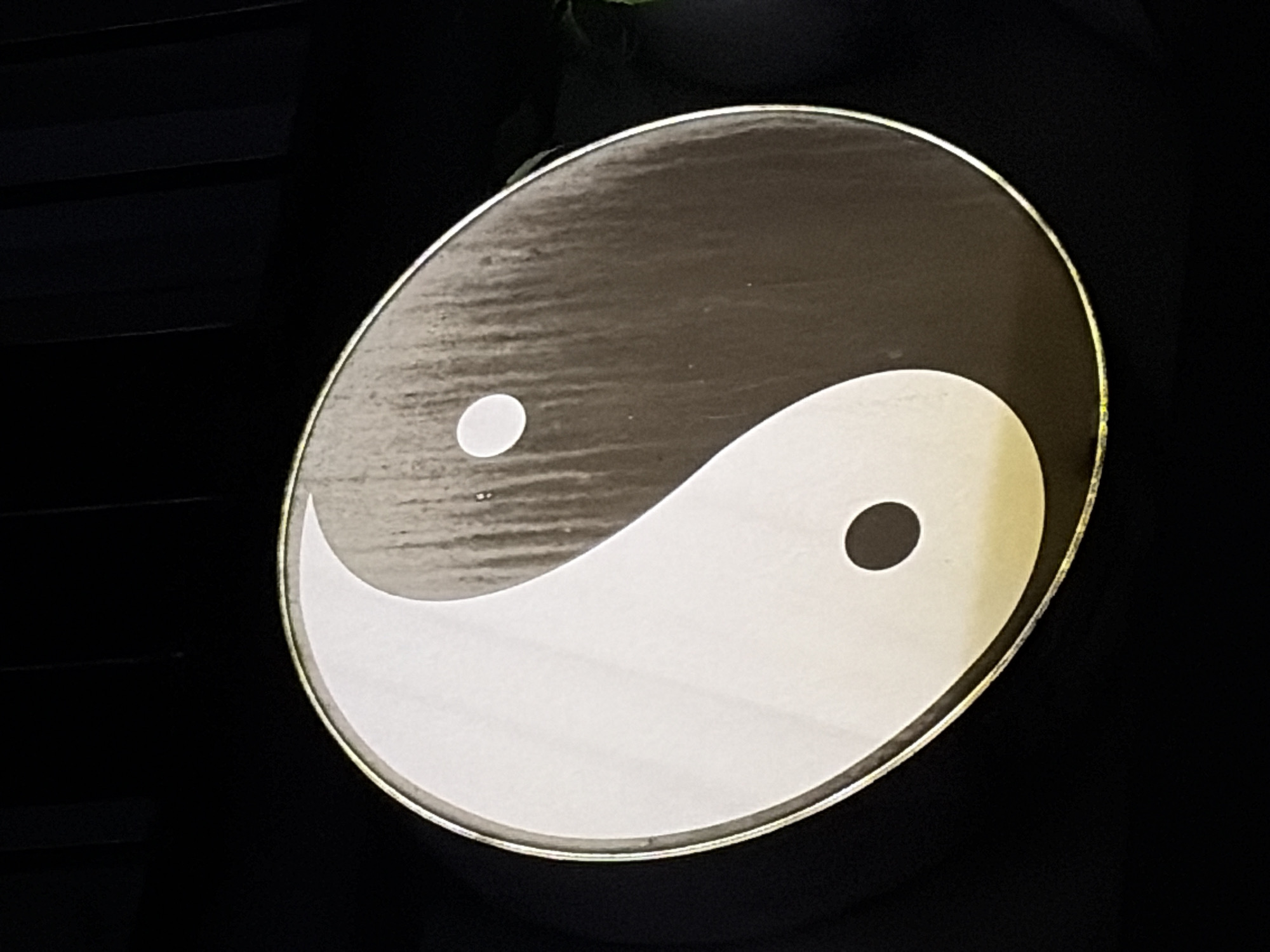 The image shows a close-up of a Yin and Yang symbol against a black background. The symbol is circular, with a gold trim around its edge. The left side of the symbol is white, and the right side is dark gray, with vertical streaks that suggest a textured or weathered surface. Each side contains a smaller circle of the opposite color: a black circle is in the white section, and a white circle is in the dark gray section. The lighting casts a slight yellow hue on the bottom portion of the symbol. The background is a plain, dark black, with some subtle vertical lines visible in the upper portion, possibly indicative of a textured wall or fabric. No people or specific city details are discernible from the image.
