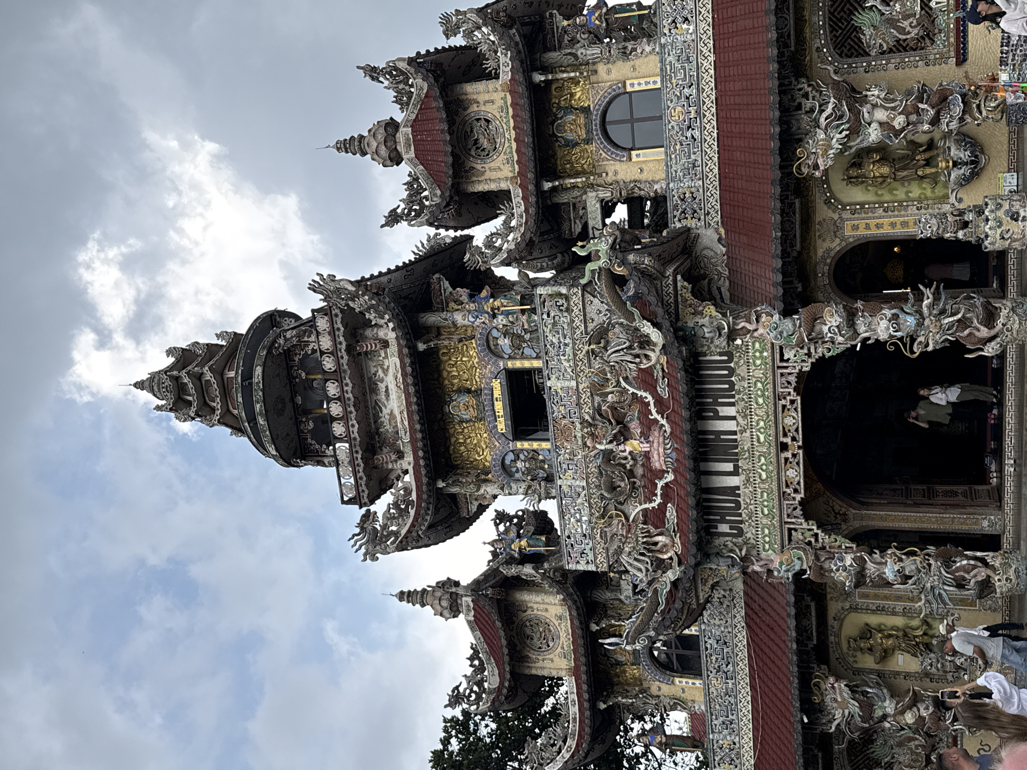This vertically-oriented image captures a section of an extraordinarily ornate multi-tiered temple, likely a pagoda, under an overcast or partly cloudy sky. The building is a spectacle of intricate architecture, adorned with countless mosaic-like decorations crafted from fragments of ceramic, glass, and pottery. Mythical creatures, prominently dragons and phoenix-like birds, are integrated into the eaves, roofs, and facades, rendered in vibrant colors and dynamic forms. Golden statues and bas-reliefs are interspersed among the colorful patterns, adding to the grandeur.
The temple features multiple levels with distinct red-tiled roofs and spire-like finials. Arched doorways and a round window are visible within the elaborate structure. On one prominent facade, the name "CHÙA LINH PHƯỚC" (Linh Phuoc Pagoda) is clearly inscribed in large white letters. Other visible text includes "禪寺雙修" (Chan Temple Double Cultivation) and "欲知前世因" (If you want to know the cause of your past life), suggesting Buddhist themes.
Several people are visible, indicating visitors or worshippers. One person stands within a dark arched entrance, while others are seen near the base of the structure, some appearing to observe or photograph the detailed artistry with their phones. The overall impression is one of immense artistic craftsmanship and spiritual significance, characteristic of Vietnamese Buddhist architecture, consistent with the location context of Xuân Thành, Vietnam. The time of day appears to be daytime, with diffused light due to the cloudy conditions.