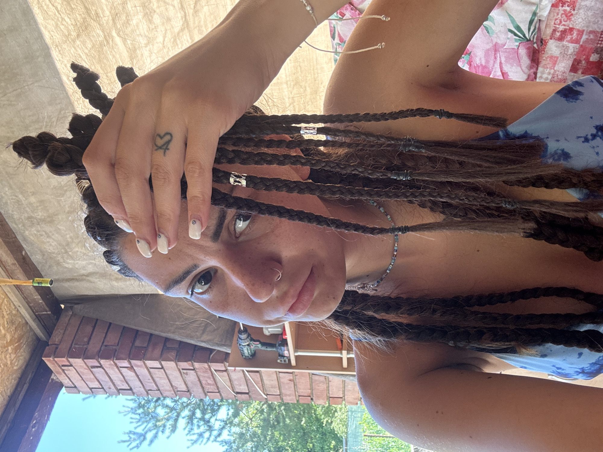 A young woman with dark, braided hair, some of which is styled in buns, is shown from the shoulders up in a selfie. She is wearing a blue and white tie-dye top and a beaded necklace. Her right hand is raised to her forehead, revealing a small tattoo on her index finger. She has a nose ring and painted nails. In the background, a brick pillar, a shelf with a power drill, and trees are visible. The scene appears to be outdoors, possibly in a backyard or workshop area. The image was captured in Belopoptsi, Bulgaria.