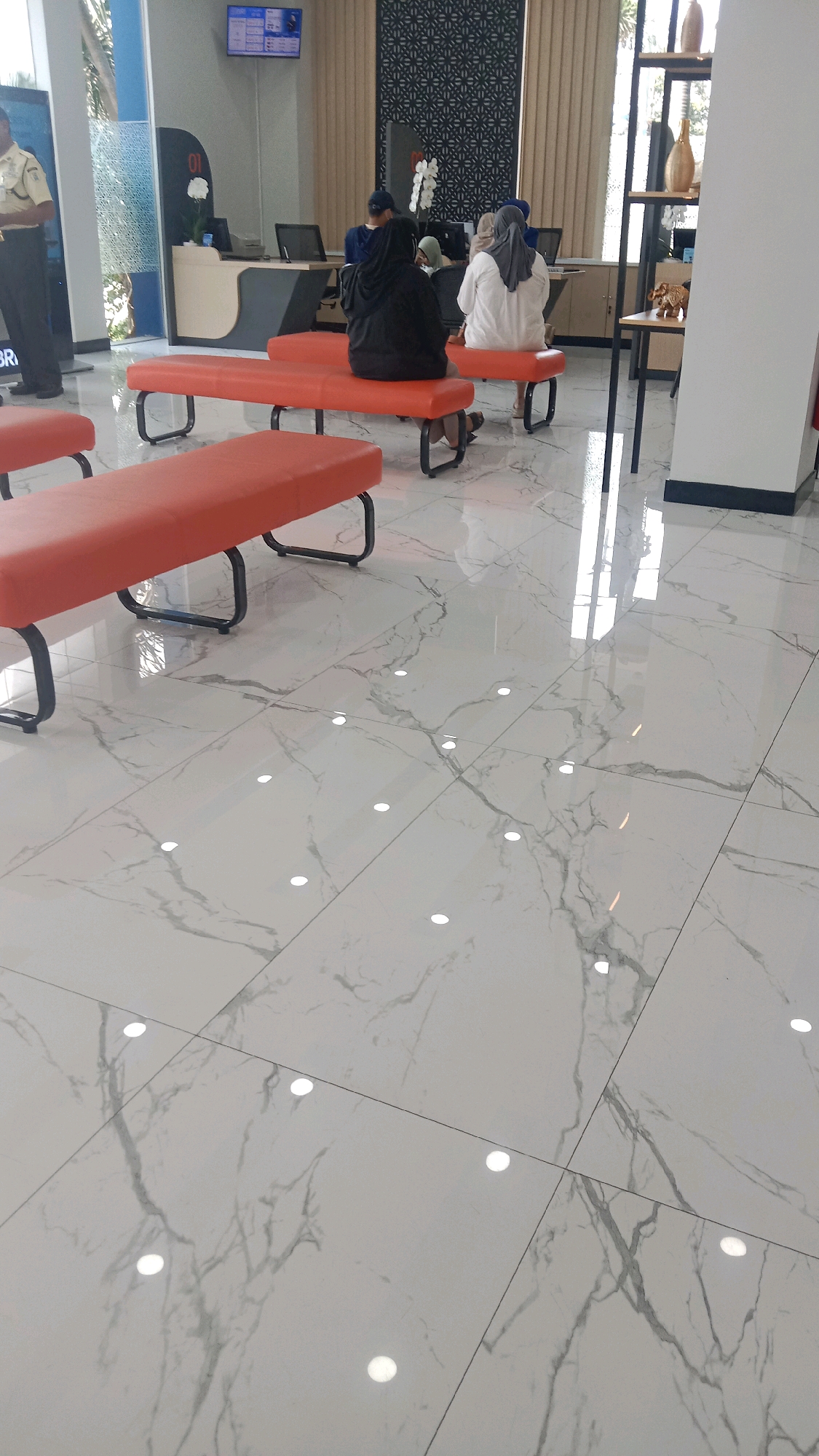 The image depicts the interior of what appears to be a bank lobby in Bekasi, Indonesia. In the foreground, three orange benches are visible, two of which are occupied by individuals. The flooring is characterized by large, white marble tiles with gray veining. In the middle ground, several people are seated on similar orange benches, facing away from the camera, towards what seems to be a service counter. Two women, each wearing a hijab, are prominently seated. One woman is wearing a black hijab and the other a white one. Another individual, possibly a man, is visible behind them wearing a blue shirt and a dark cap, facing the counter. A security guard in a tan uniform stands to the left of the frame, facing left. In the background, a service counter with computers and a decorative screen are visible. The overall environment suggests a clean, modern, and organized public space.