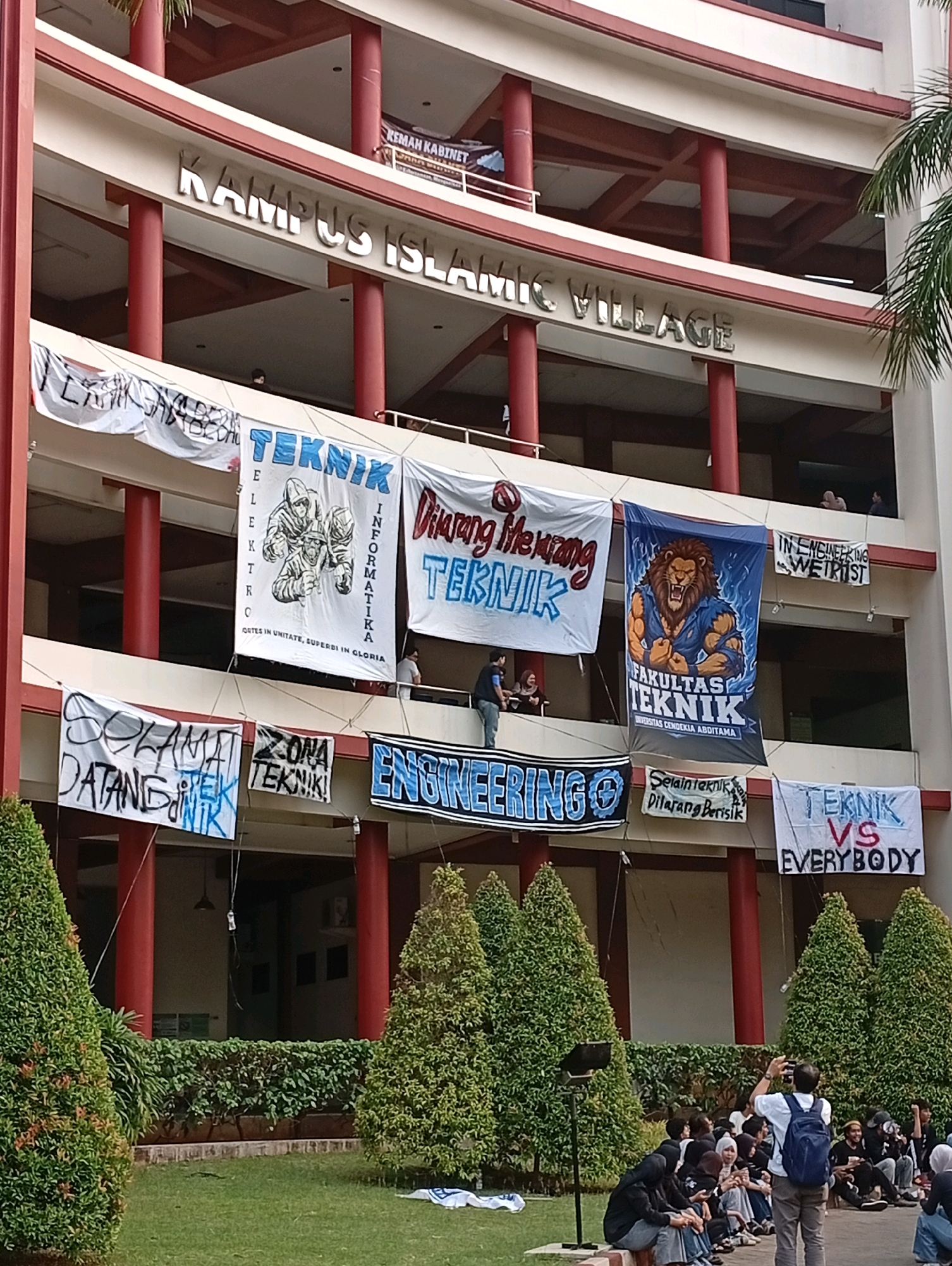 The image captures a facade of a multi-story building with a sign that reads "KAMPUS ISLAMIC VILLAGE" in large, metallic letters. Red pillars support the building's balconies, and several large banners hang from the upper levels. One banner features a roaring lion in a blue background and text that translates to "FACULTY OF ENGINEERING". Another banner displays three figures in hoodies and the word "TEKNIK" (Engineering). Other banners have text like "SELAMAT DATANG TEKNIK" (Welcome Engineering), "ZONA TEKNIK" (Engineering Zone), "ENGINEERING", and "TEKNIK VS EVERYBODY".
Below the building, a group of students, mostly wearing black, are seated on the grass and a low wall, facing the building. Some are looking at their phones, while others are engaged in conversation. One person in the foreground, wearing a blue backpack and grey pants, is standing and taking a photograph with a phone. The environment includes manicured green trees and a lawn. The scene appears to be at an educational institution in Kelapa Dua, Indonesia, likely during an event or orientation for engineering students.