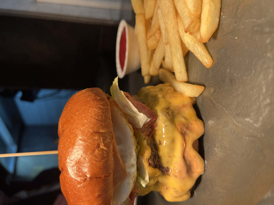 A large, double-patty burger, secured by a wooden skewer, is presented on a dark, textured surface. The burger features a golden-brown brioche bun, two beef patties covered in copious amounts of melted yellow cheese, multiple strips of cooked bacon, shredded green lettuce, and a fried egg with a solid white and partially obscured yolk. To the right of the burger, a pile of golden-brown french fries is arranged, with a small white ramekin containing red ketchup positioned behind them. The background is dark and out of focus, indicating an indoor setting, possibly a commercial kitchen or restaurant environment. A blurry light blue area is visible in the upper right background. No individuals or specific actions are depicted. The specific location within Budva, Montenegro, is not discernible from the visual information provided.