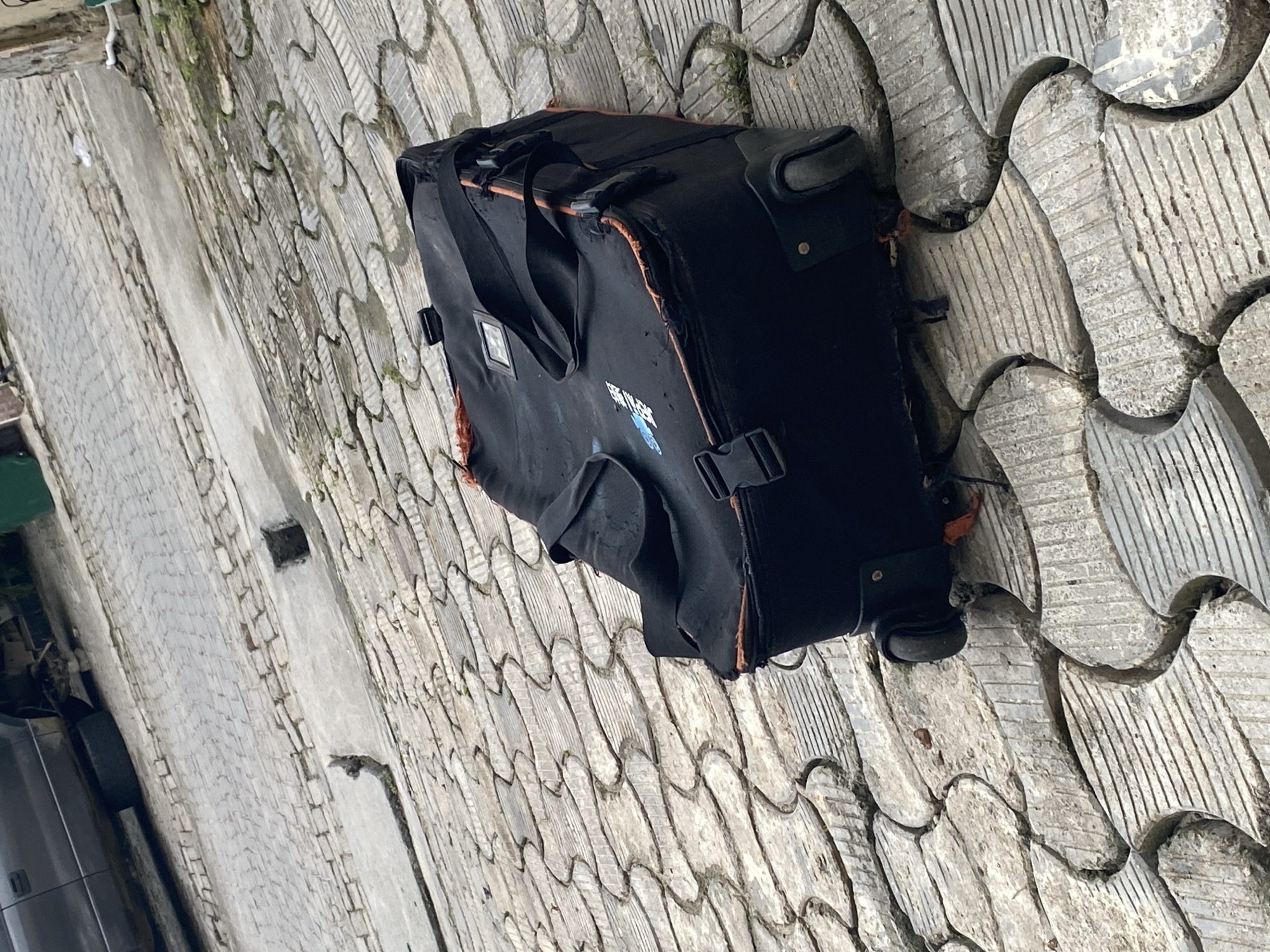 A black, soft-sided wheeled bag, appearing to be a duffel or suitcase, rests prominently on patterned grey pavement. The bag shows significant wear, with frayed edges exposing an orange-brown lining. It features a retractable handle, wheels, multiple black straps, and plastic buckles. A small, transparent identification pocket is visible on one side. On the main body of the bag, a partially visible blue graphic displays text that appears to read "EXTREME GEAR," accompanied by a stylized blue symbol, possibly an animal or abstract design.
The pavement beneath the bag is constructed from interlocking concrete pavers, each featuring a distinct wavy or scalloped shape, some with fine parallel grooves for texture. Green moss or small plants grow in the gaps between these pavers, particularly on the left side of the image. To the left of the patterned pavement, the ground transitions to a strip of smoother, lighter grey concrete, and further left, the front section of a dark-colored vehicle is partially visible. The scene is outdoors, likely during daytime, under diffused light as indicated by the lack of strong shadows. No people are present, and the bag is stationary, suggesting it is either awaiting transport or has been temporarily placed.