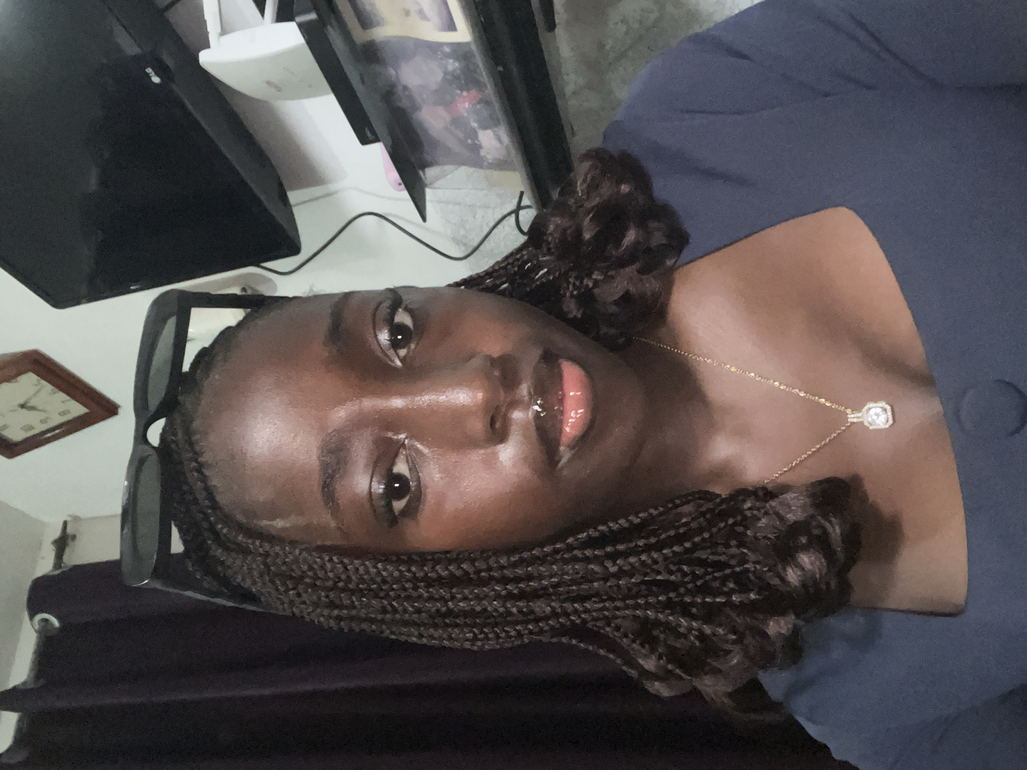 A medium shot, rotated ninety degrees, shows a dark-skinned woman in a blue garment with a gold chain necklace that has a square pendant. Her hair is braided and styled around her face. Sunglasses are placed on her head. The woman looks into the camera.
In the background, there is a television, a white router, a picture frame, and a clock. The exact location within Abuja, Nigeria, cannot be determined from the available information.