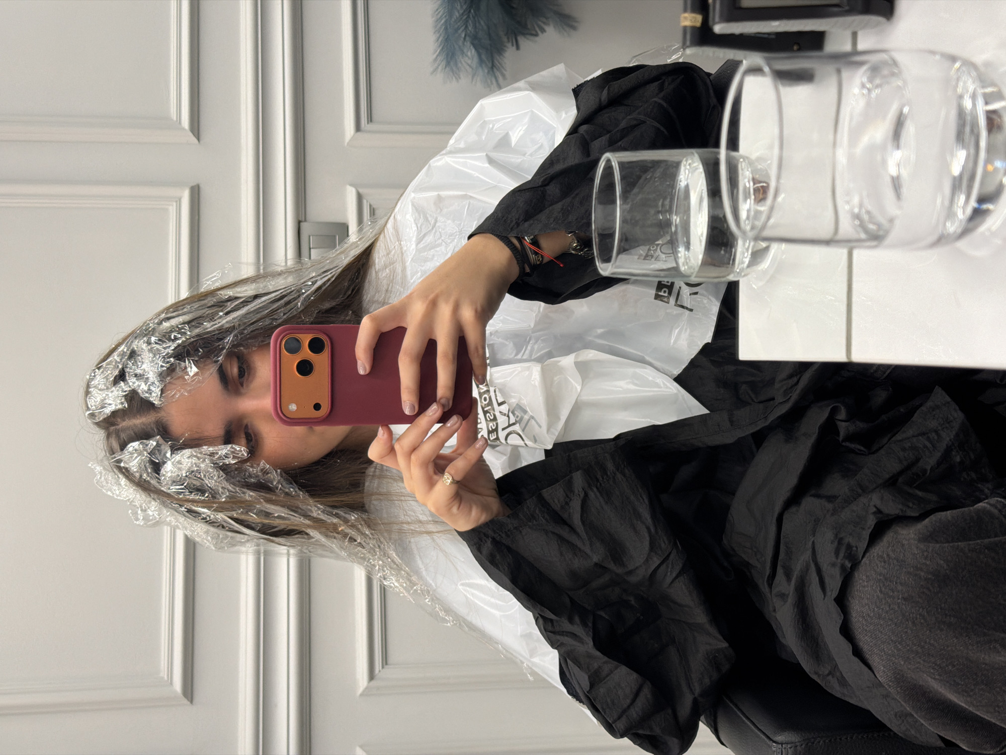 A young woman is captured in a mirror selfie, in an indoor setting that appears to be a hair salon or a well-appointed home. She is undergoing a hair treatment, with sections of her long brown hair wrapped in aluminum foil and clear plastic film, suggesting highlights or coloring. She is draped in a black smock or cape for protection, over which a crinkled white plastic sheet is also visible. Her left hand holds a red/maroon smartphone with a triple-lens camera, used to take the reflection photo, partially obscuring her face, though her right eye and nose are visible.
Her left wrist is adorned with multiple bracelets, including red and black string bands and a metallic one. A ring is visible on her right ring finger, and her fingernails are painted a light, neutral color.
The background features a white paneled door with classic detailing, indicating a neat and possibly upscale environment. To the right, a white table surface holds two clear drinking glasses, one larger and one smaller, both appearing empty or containing clear liquid. A small, light blue artificial plant or decoration is discernible in the upper background. Part of a black, possibly leather, chair is seen at the bottom right. Reflected on a white plastic bag in the background, text resembling "KOISS∃ᴚ" (a reversed word, possibly a brand like "RESISTO" or similar) is visible. The atmosphere is calm and focused on the beauty treatment.