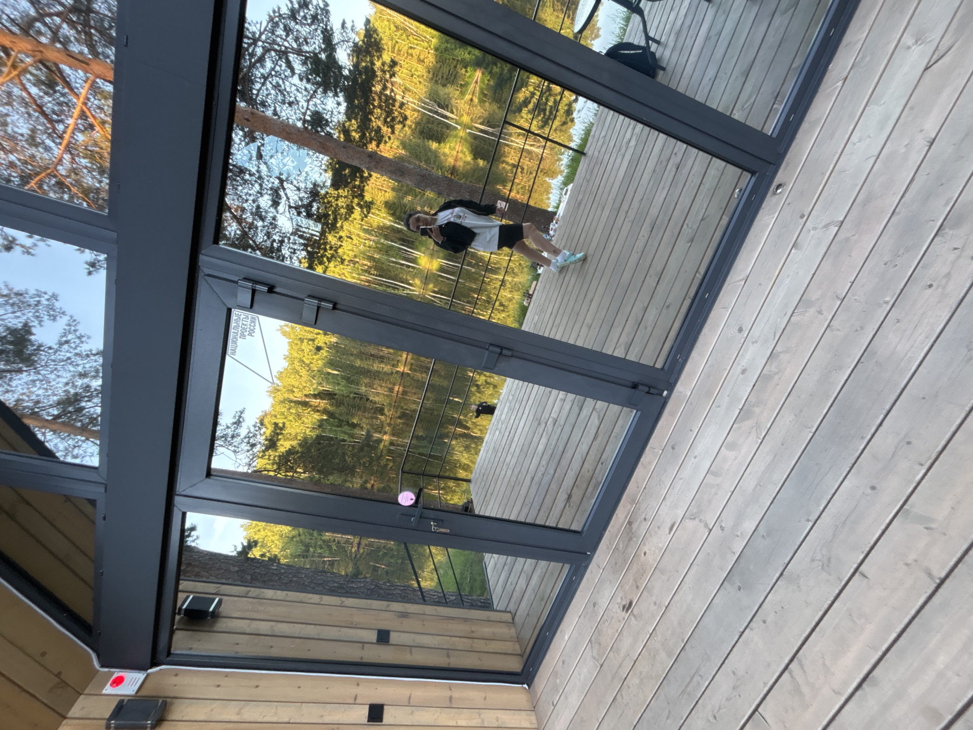 A person, appearing female, dressed in a white t-shirt, black jacket, black cycling shorts, and light blue athletic shoes, is reflected in large glass doors and windows of a modern building in Mishkino, Russia. The individual is positioned on a weathered wooden deck, holding a smartphone to capture the reflection.
The building features dark gray frames for the floor-to-ceiling glass panels, with a light-toned wooden facade on the left. A prominent white sticker with black Cyrillic text, "НАЦИОНАЛЬНЫЕ ПРОЕКТЫ РОССИИ" (NATIONAL PROJECTS OF RUSSIA), is affixed to an upper glass panel. A small pink circular sticker is on the handle of the central door, which includes a keyhole. Exterior elements visible on the wooden facade include a square black electrical box, a red light sensor, and white conduit. Further inside the reflection on the left, a black speaker or similar device is mounted on the wooden wall.
The reflection reveals a black metal railing separating the deck from a calm body of water, likely a lake or river, which stretches towards a dense forest on the opposite bank. The forest exhibits a mix of green foliage and lighter-barked trees, illuminated by what appears to be late afternoon or early morning sunlight. The sky, visible through the upper glass, is clear blue. A dark backpack and part of a table are visible on the deck in the reflection on the right side. The wooden deck itself shows signs of wear and some minor discoloration.