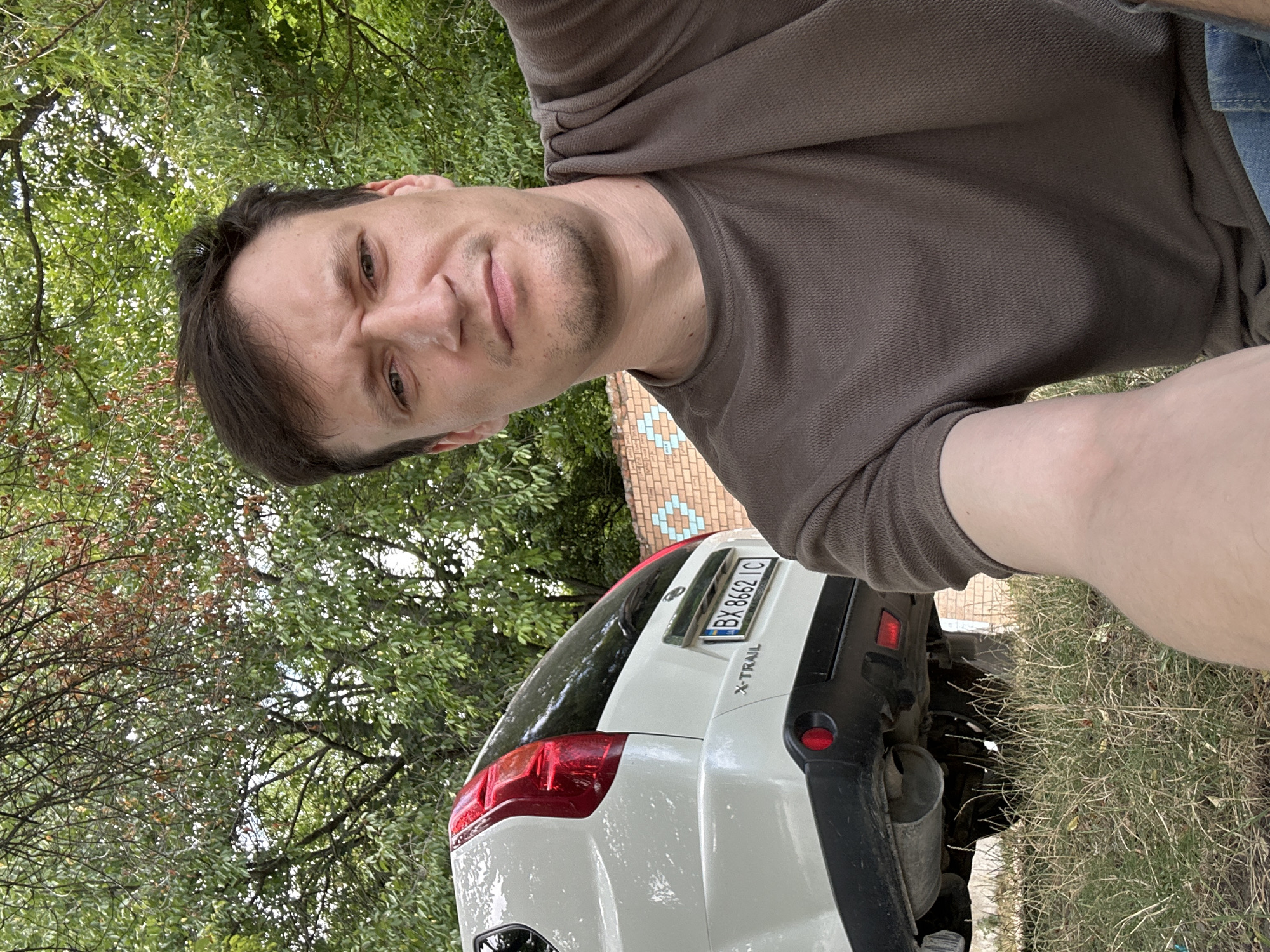 A man with a brown shirt and jeans is taking a selfie outdoors. Behind him, to the left, is the rear of a white Nissan X-Trail car. The car has a license plate that reads "BX 8662 IC". The background consists of lush green trees and a brick wall with a blue heart pattern. The ground is covered with dry grass and some fallen leaves. The man appears to be in a casual setting, possibly a yard or park, in Makiv, Ukraine.