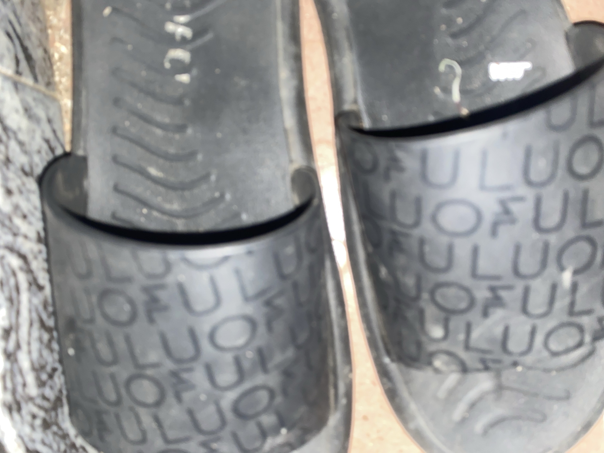 A close-up, overhead shot displays two black slide sandals on a textured, beige-brown tiled surface. The sandals are positioned side-by-side, with the top sandal slightly angled to the left and the bottom sandal parallel to the frame. Both sandals have a repeating embossed pattern of the letters "U" and "L" throughout their footbeds. The right sandal of the pair exhibits a faint "FC" logo embossed on its strap. The background consists of a speckled, gray and white tiled surface that partially covers the top of the sandals. The scene is devoid of people or any discernible actions.
