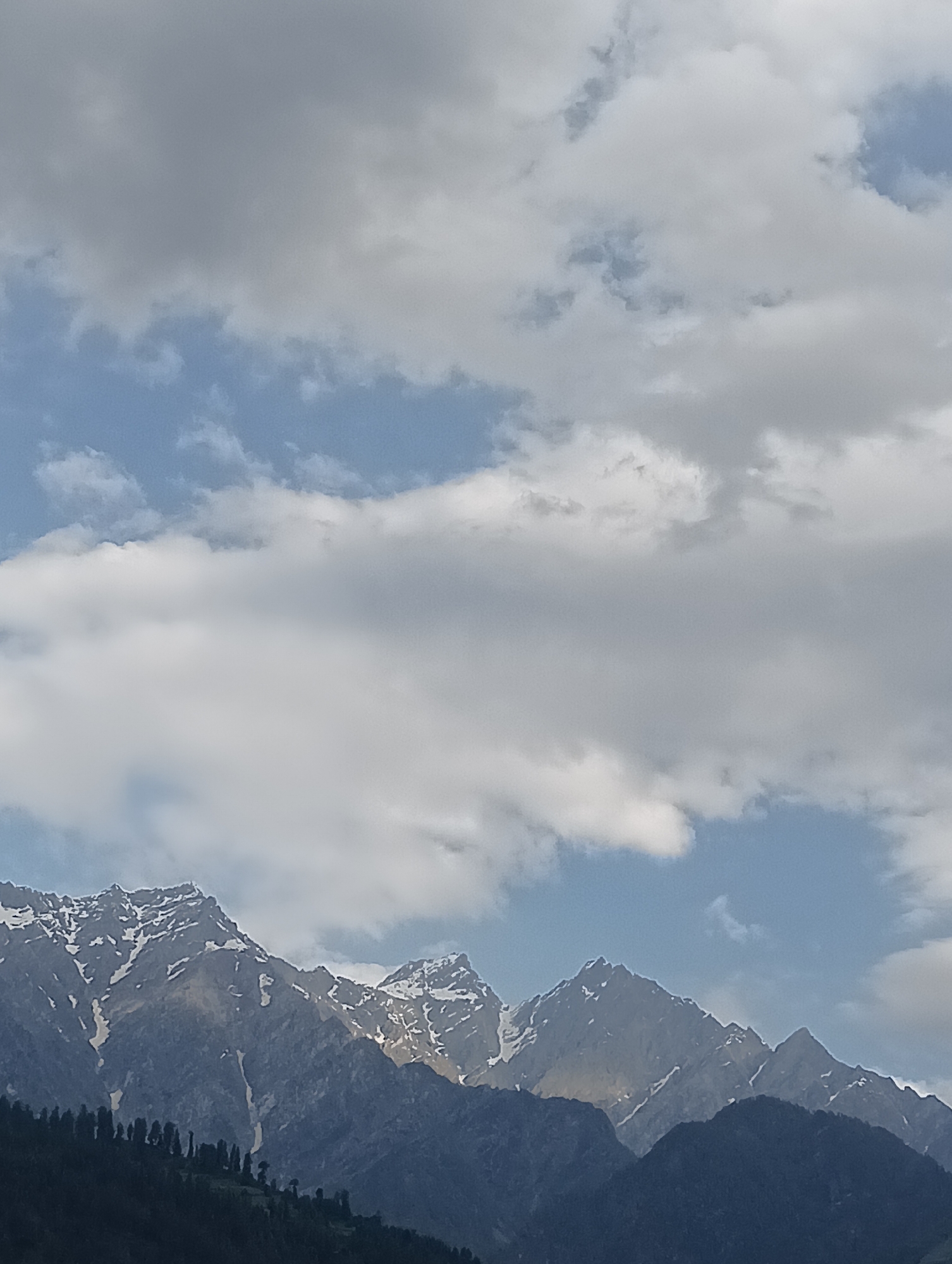 A photograph captures a majestic view of snow-capped mountains under a cloudy sky. The foreground features a dark, densely forested slope. The mountains themselves are rugged and imposing, with patches of white snow accentuating their peaks and ridges. The sky is a mix of blue and gray clouds, suggesting a dynamic weather pattern. The overall impression is one of natural beauty and grandeur, characteristic of the mountainous terrain found in and around Vashisht, India.