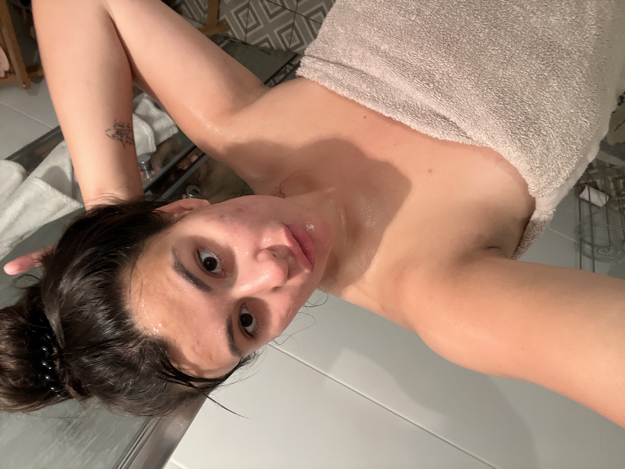 This is a portrait-oriented, medium-shot, selfie-style photograph taken in a bathroom. The subject, a young woman with her hair pulled up in a messy bun, is looking at the camera. She is wearing a light brown towel wrapped around her chest. Her skin appears wet and shiny, suggesting she has recently showered. There are some blemishes and redness on her face. The background shows a tiled wall and a shower curtain, indicating the location is likely a residential bathroom. The lighting is bright and even. No specific landmarks of Kiryat Bialik, Israel, are visible in the image.