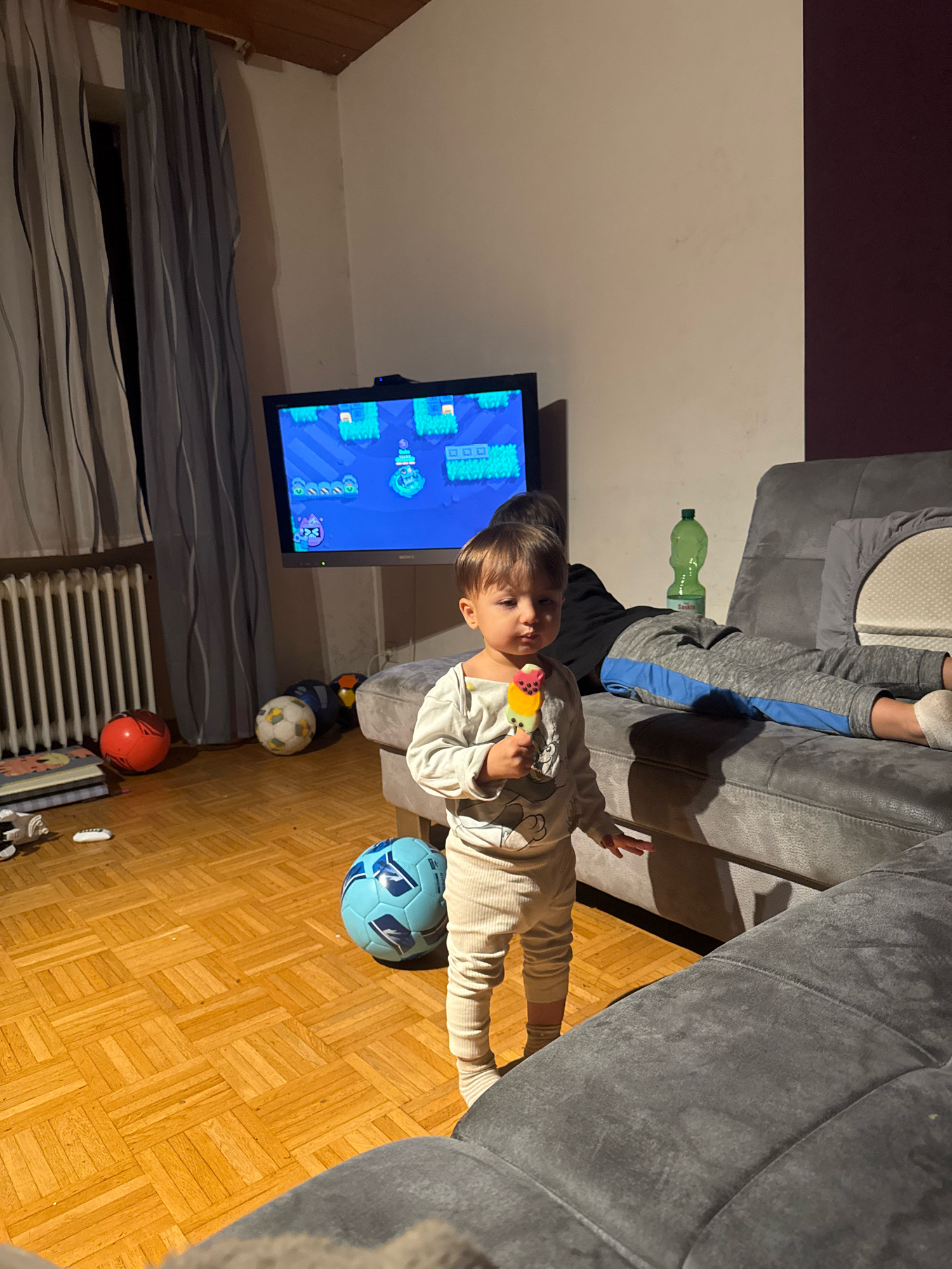 In a room with light-colored wooden flooring, a young child stands in the foreground, holding an ice cream cone. The child is wearing a light-colored long-sleeved shirt with a graphic on it and cream-colored pants. The child's feet are bare or wearing light socks. Behind the child, a grey sofa is visible, with another person, possibly a teenager or adult, lying on their stomach on the sofa. This person is wearing a black t-shirt and grey pants with blue stripes. A green plastic bottle stands on the sofa. A television screen displays a video game with a pixelated landscape. Several soccer balls are scattered on the floor: a red one, a white and black one, and a blue and white one. To the left of the television, a radiator is visible behind grey and white striped curtains. The scene appears to be an indoor living space.